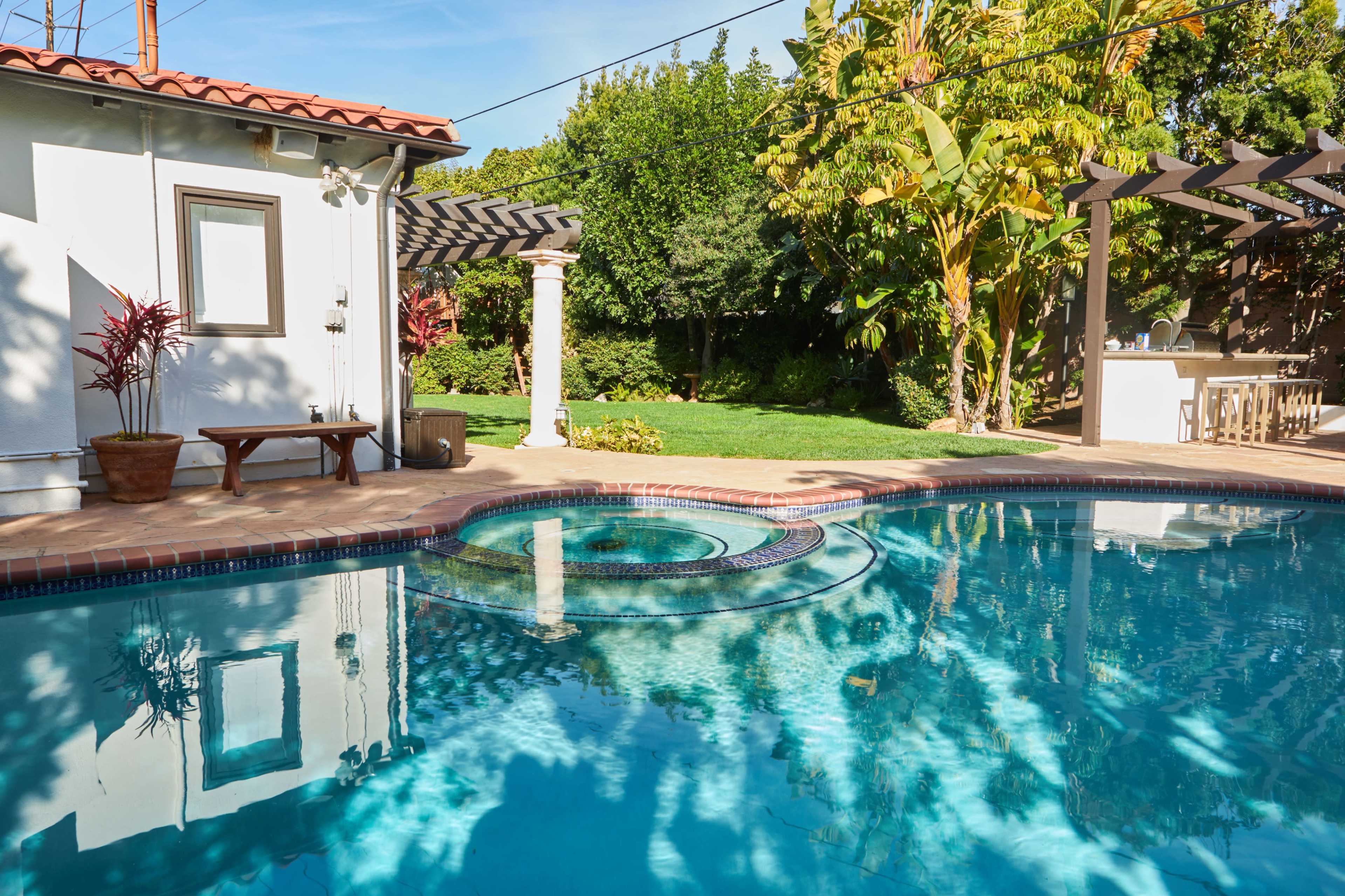 The image shows a swimming pool with a connected hot tub, surrounded by landscaped greenery and a patio with outdoor seating.