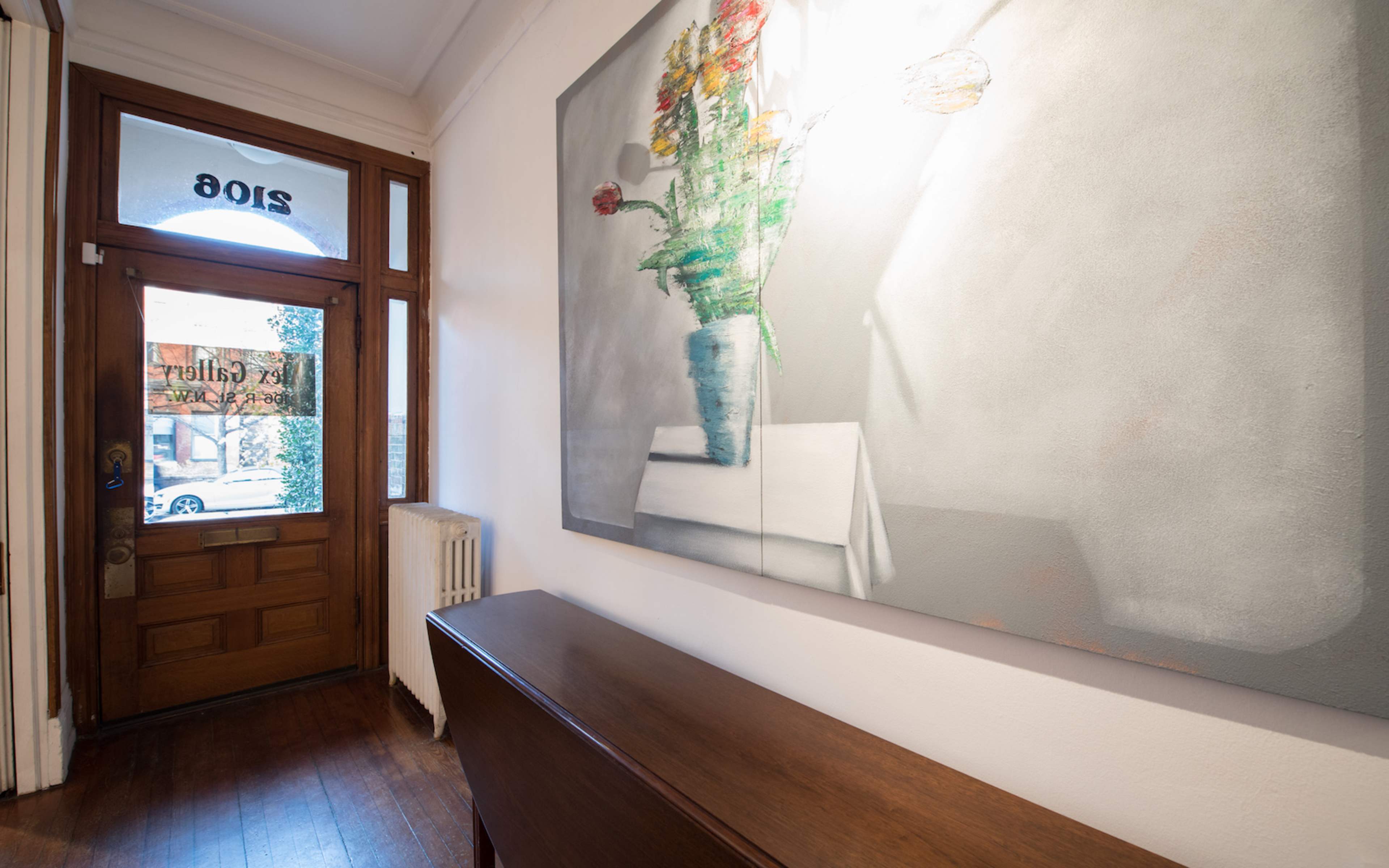 An entrance hallway featuring a wooden console table, a large flower painting on the wall, and an open door leading to the outside.