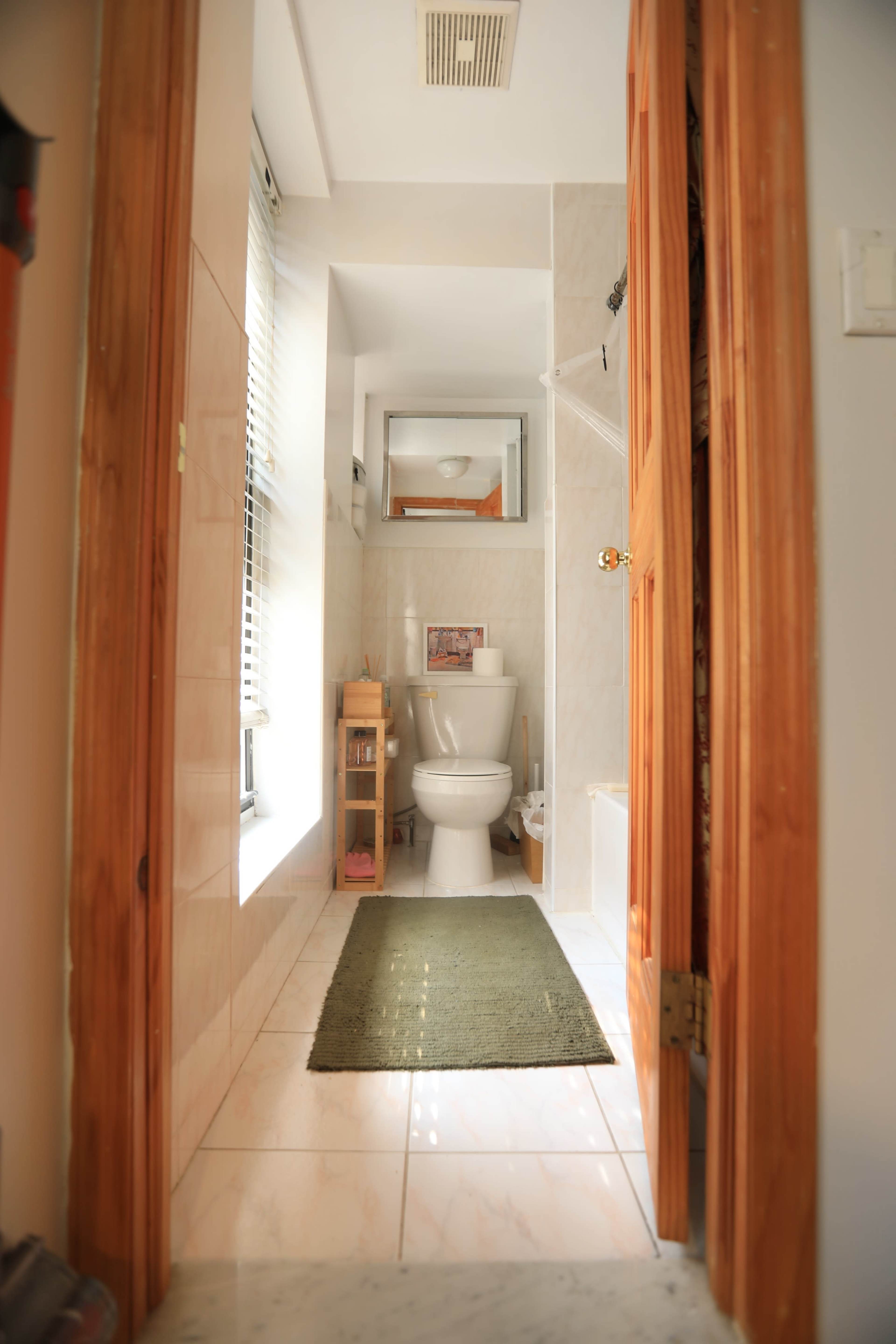 A bathroom with a toilet, a small wooden shelf, and a green rug, viewed from an open doorway.