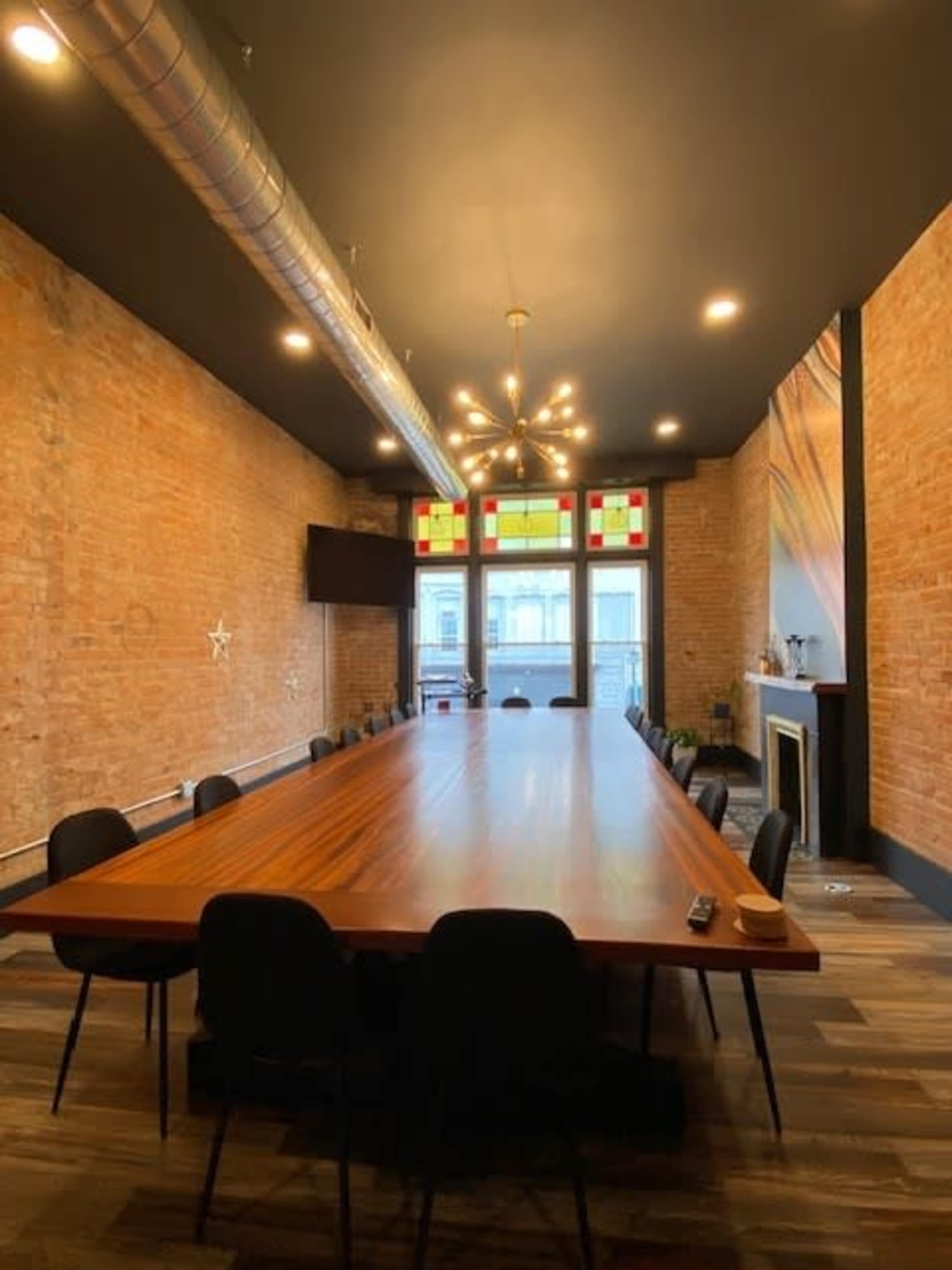 A spacious conference room with a long wooden table, surrounded by black chairs, exposed brick walls, and a large window featuring stained glass.