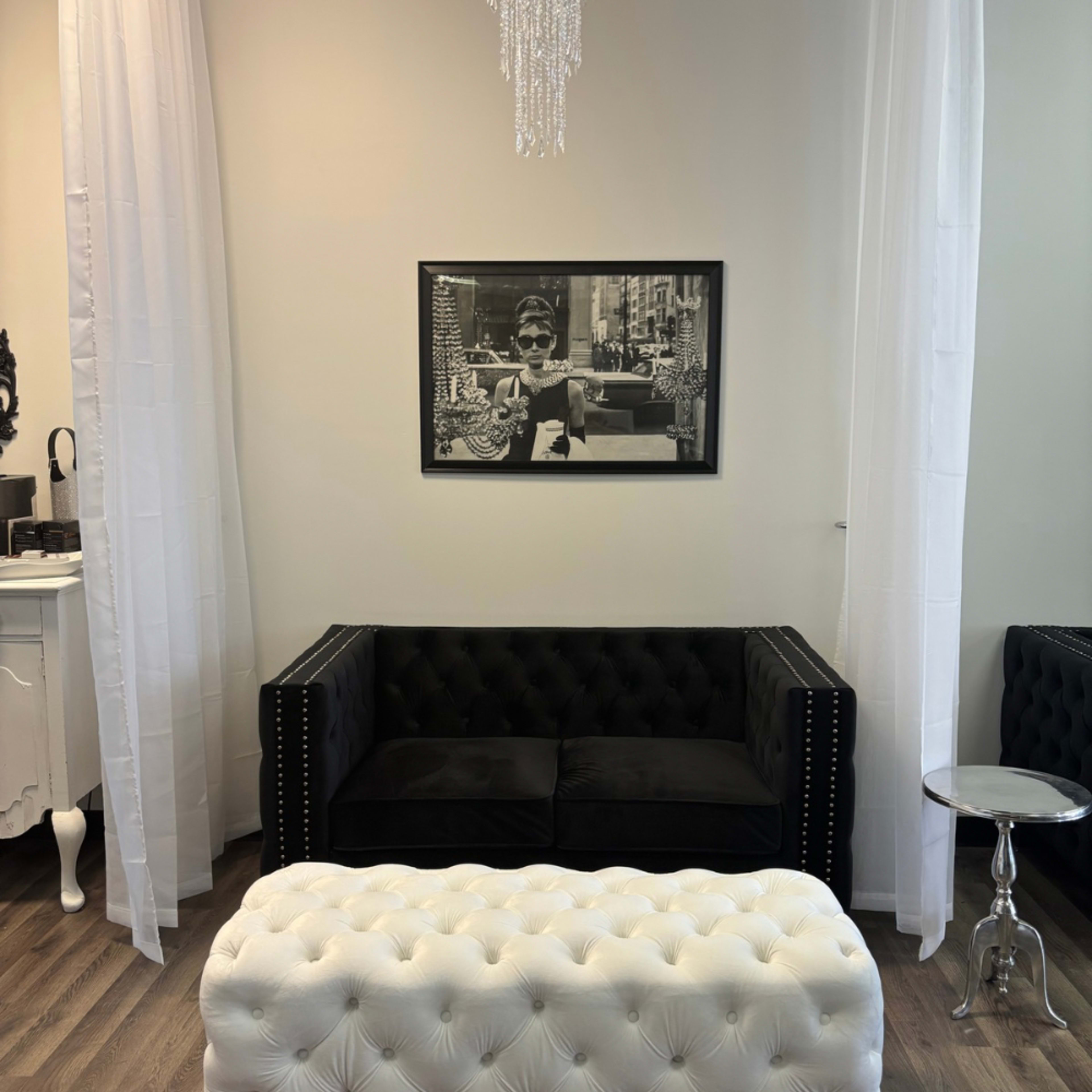 The image shows a minimalist interior with a black tufted couch, a white tufted ottoman, and a framed black-and-white photograph on the wall.