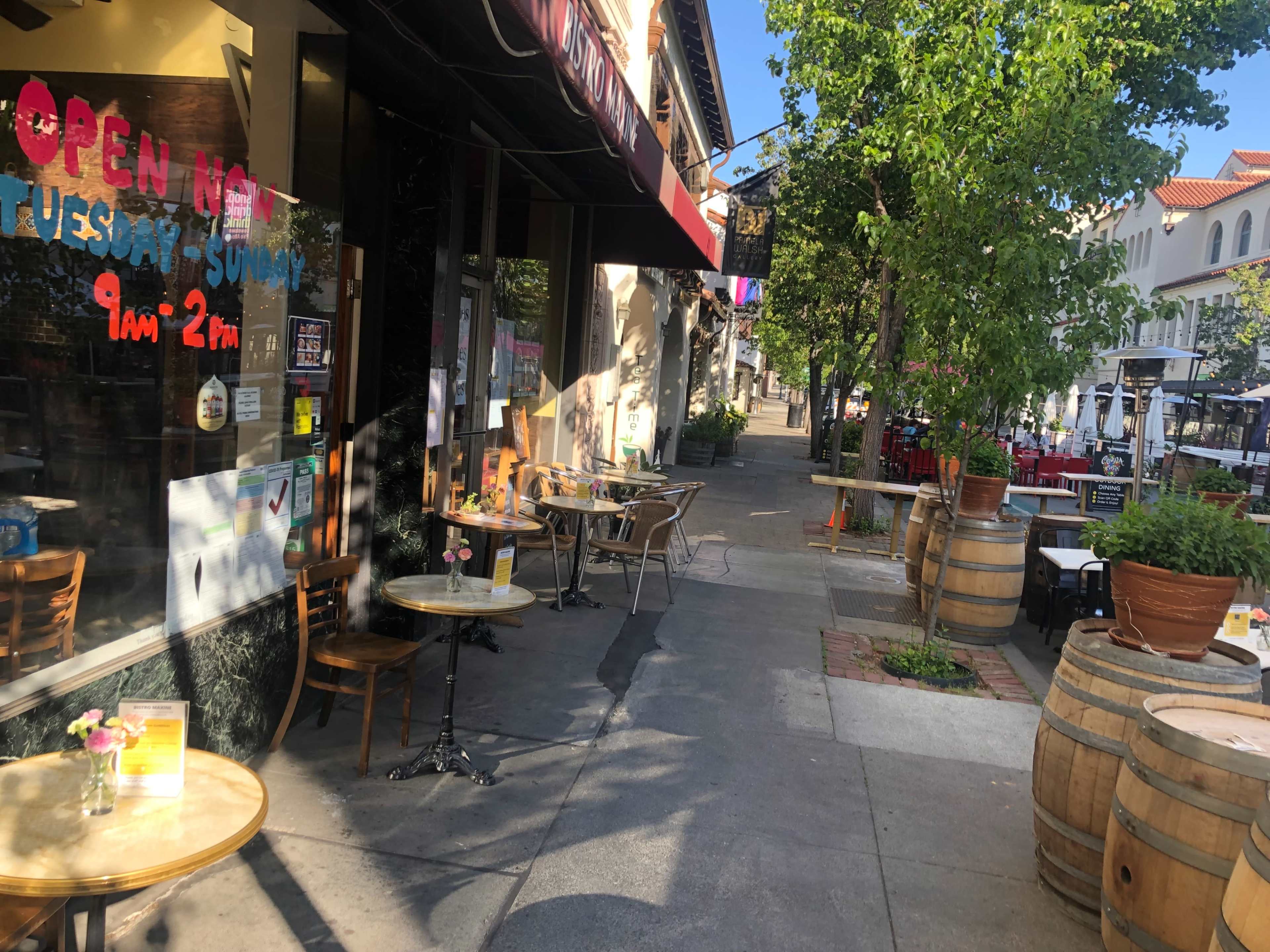 A sidewalk café with small tables and chairs is set up outside a restaurant, featuring a sign that indicates its open hours.