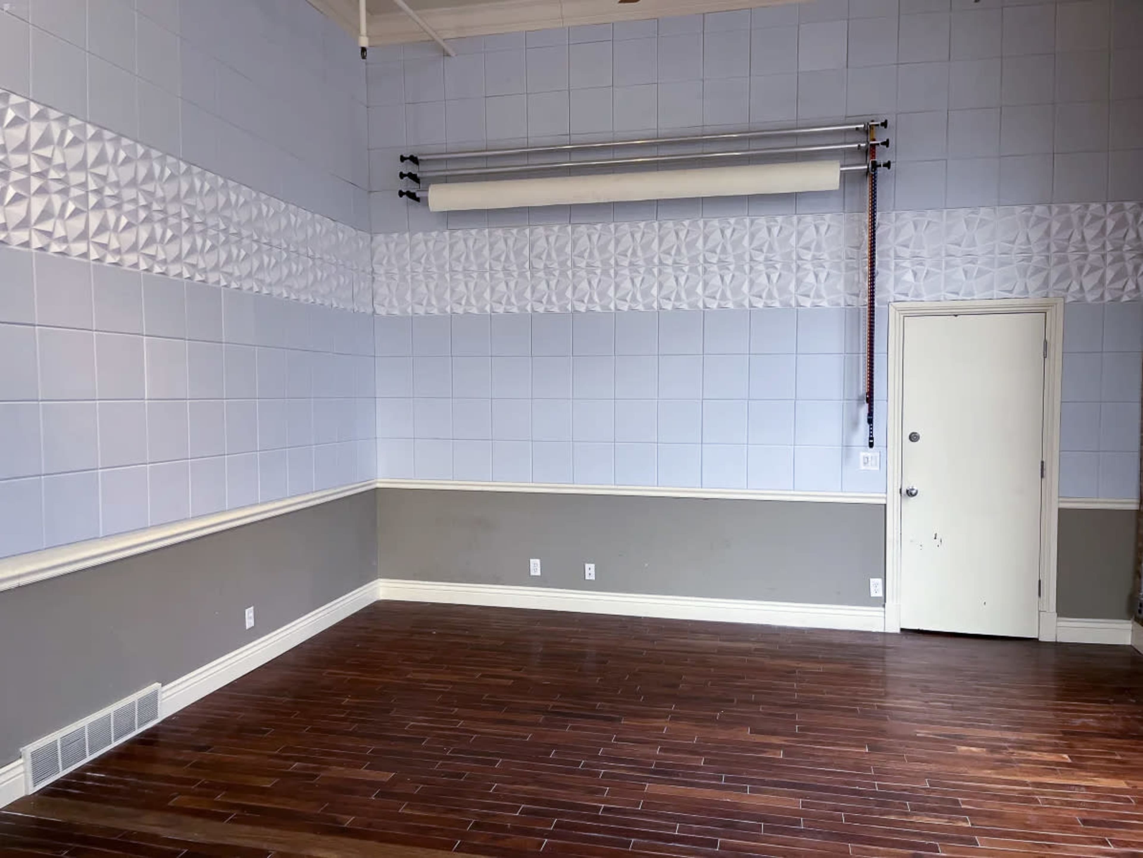 An empty room with light blue tiled walls, a decorative white panel near the ceiling, and dark wooden flooring.