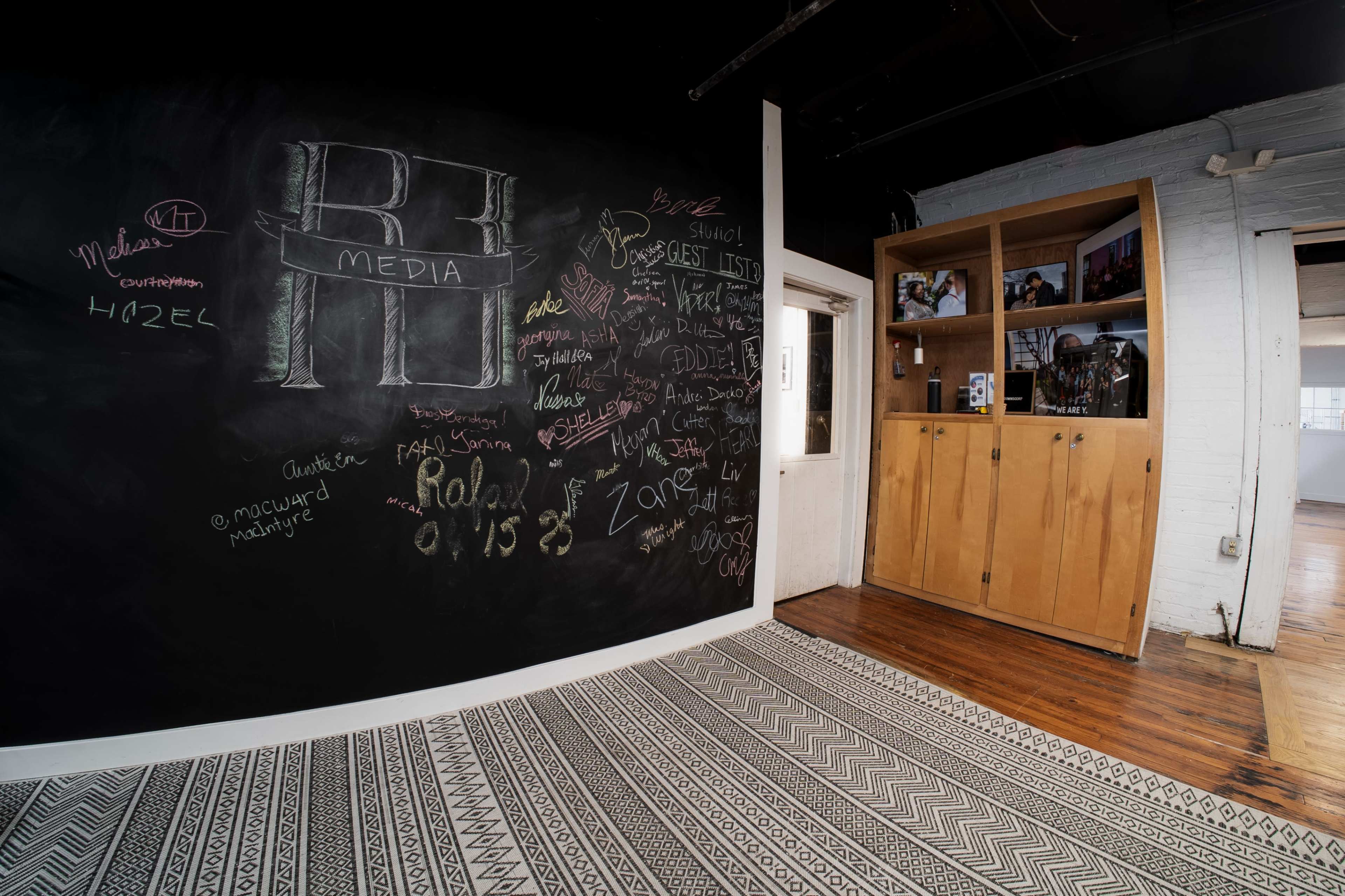 The image shows a room with a large black chalkboard wall featuring various writings and drawings, along with wooden cabinets and a patterned floor.