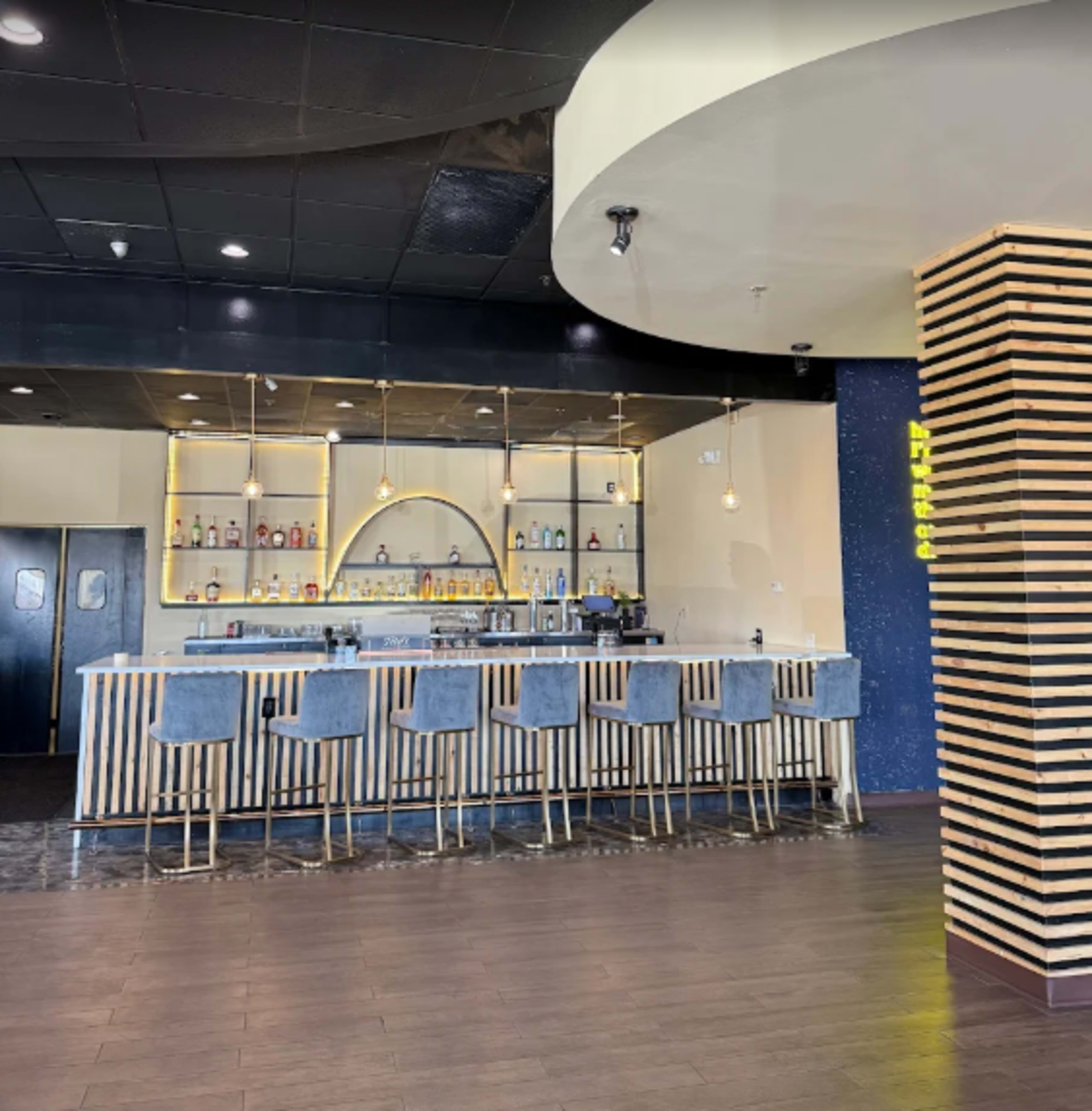 The image shows a modern bar area with a curved counter, high stools, and shelves displaying various bottles of alcohol.