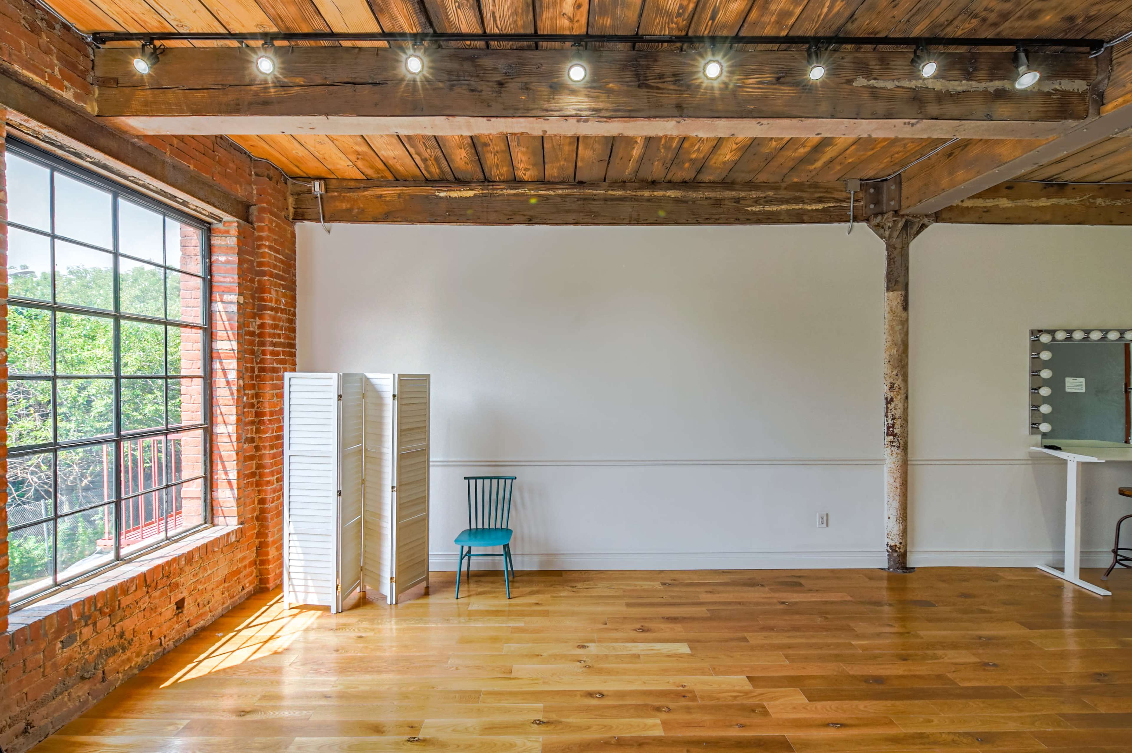 The image shows a spacious room with exposed brick walls, wooden beams, and a large window, featuring a blue chair next to a white folding screen and a bar counter.