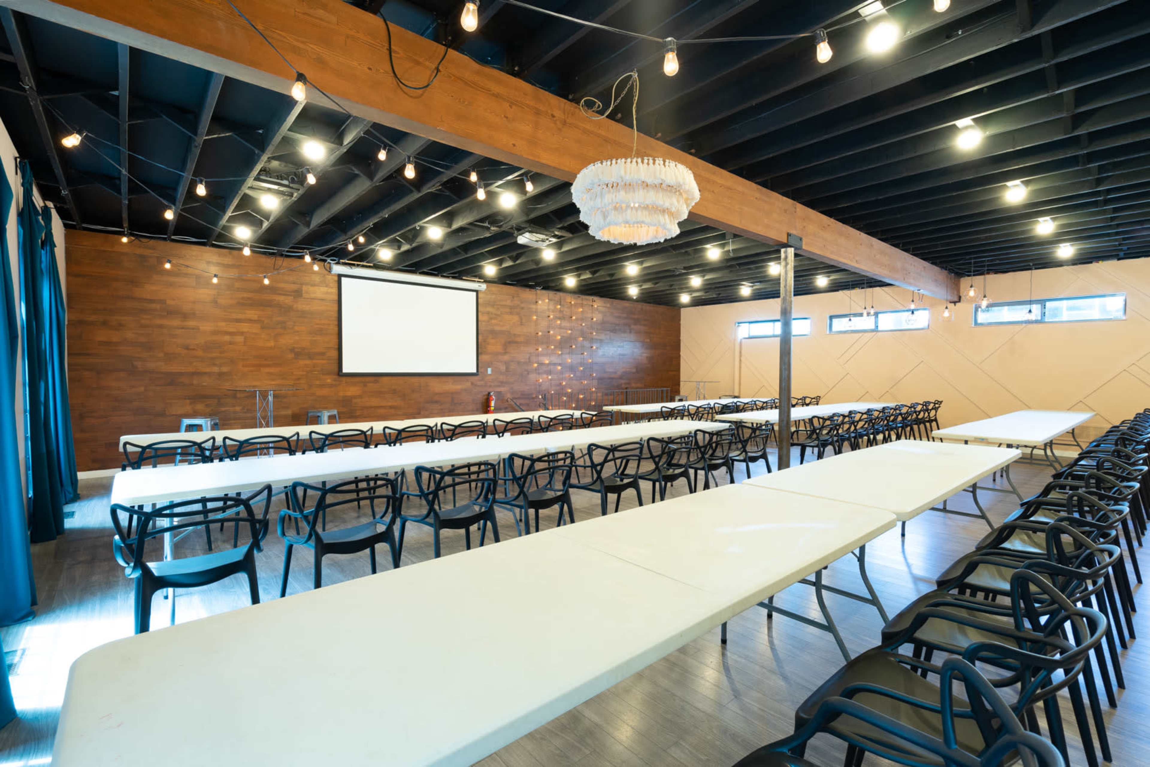 A spacious event room features multiple long tables set up in rows, with a large projector screen on one wall and string lights hanging from the ceiling.