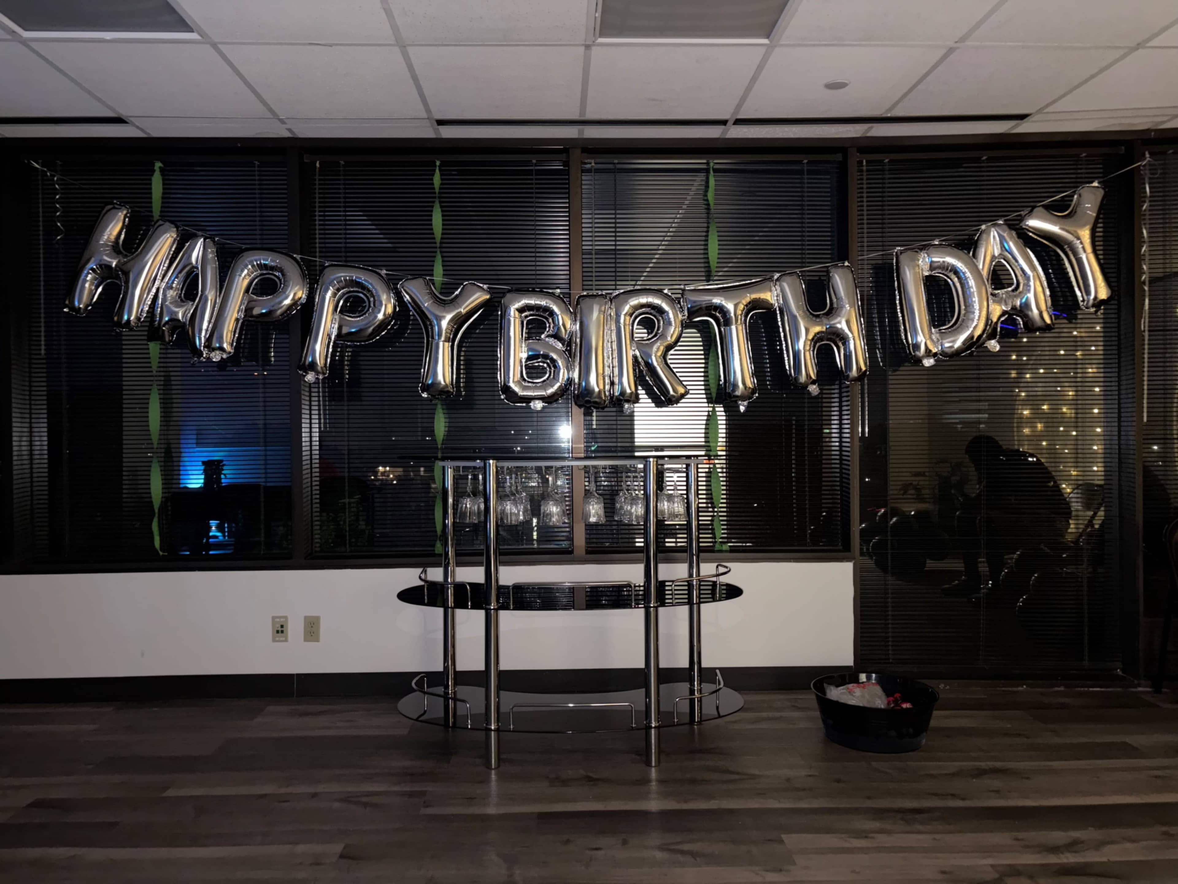 A "Happy Birthday" banner made of metallic balloons hangs over a table in a dimly lit room with large windows.