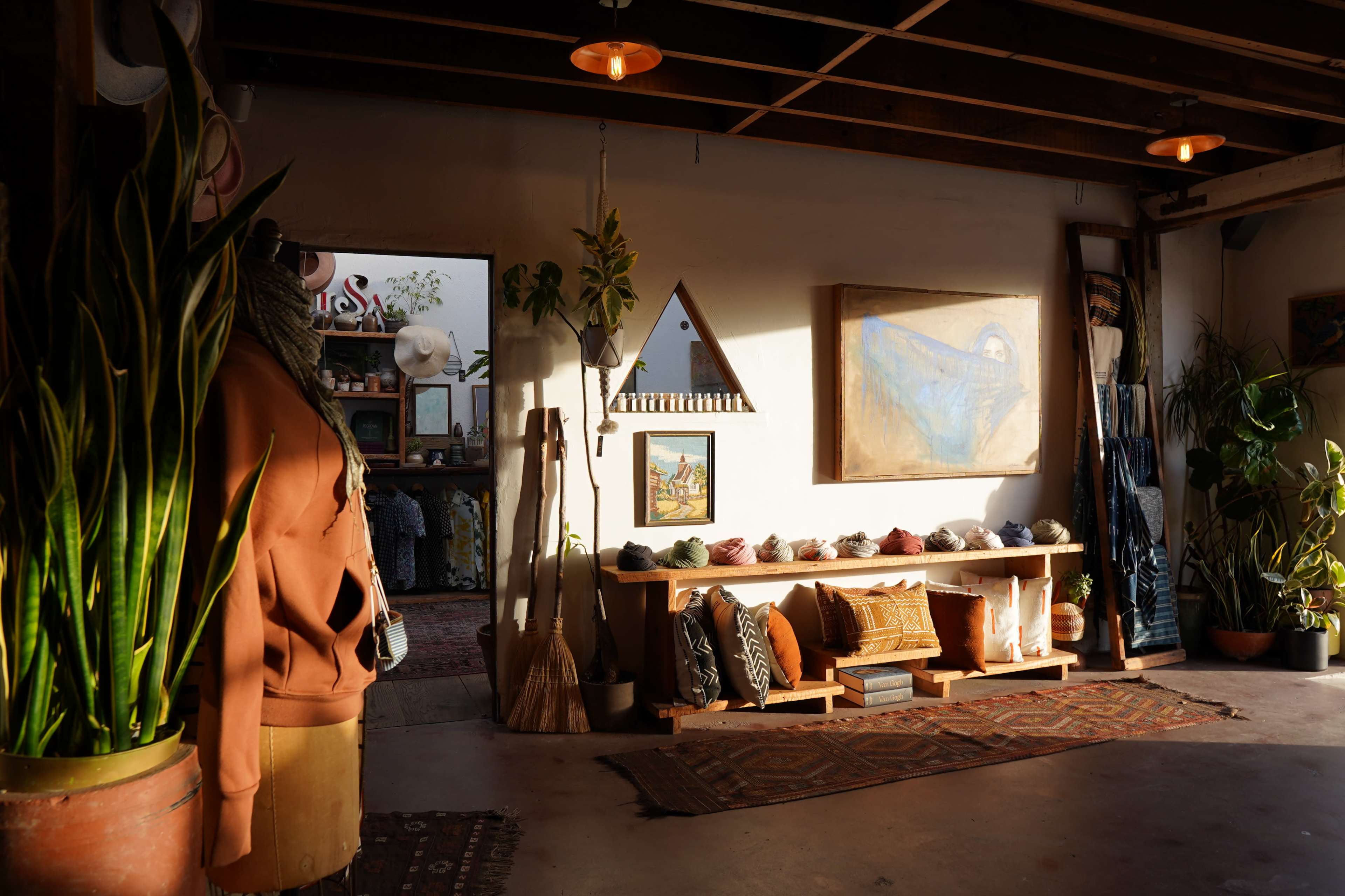 The image depicts a cozy, sunlit interior of a store featuring clothing, decorative items, and potted plants.