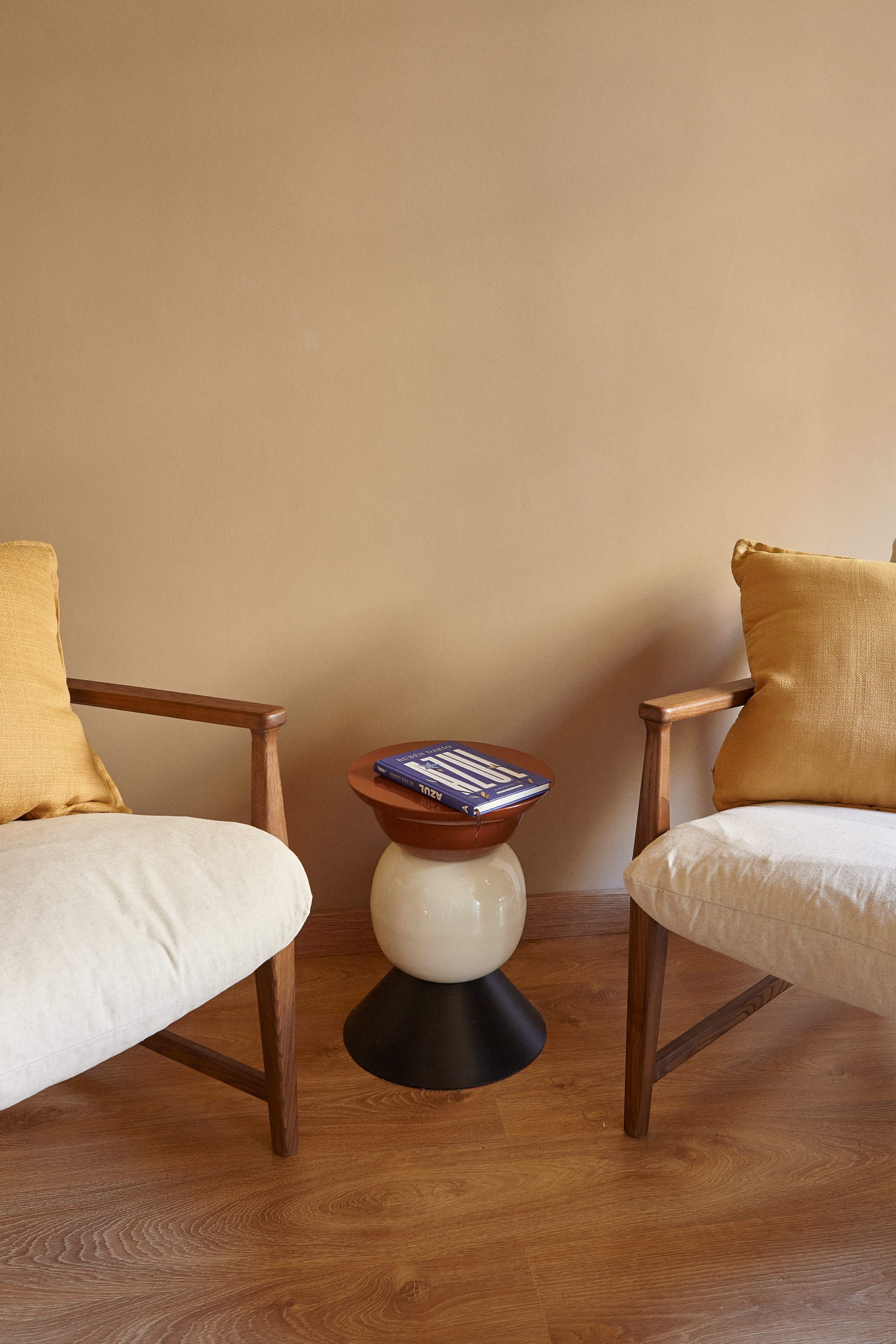 Two wooden chairs with cushions are positioned around a small, round side table on a wooden floor against a tan wall.