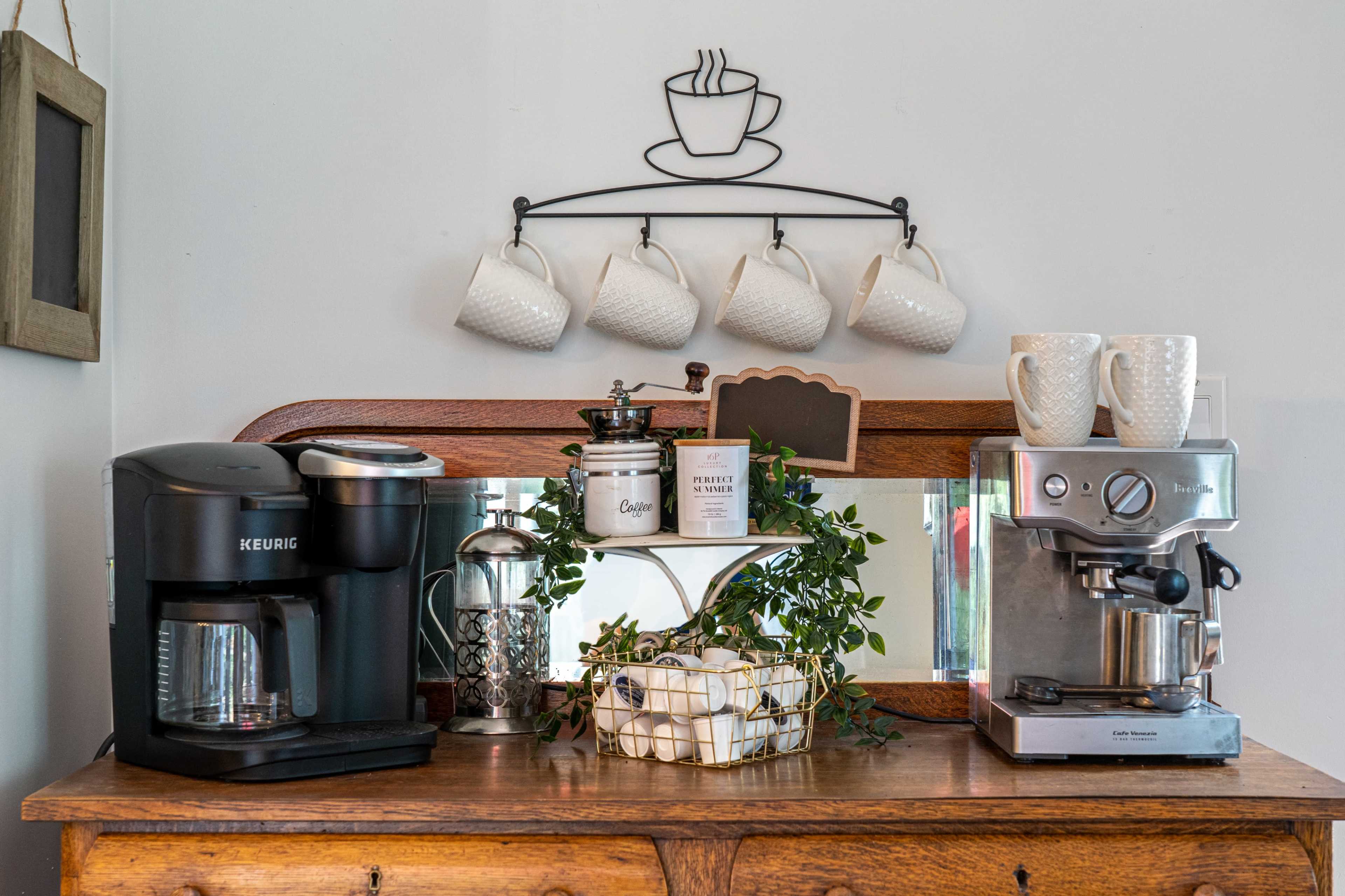A wooden countertop displays a coffee maker, an espresso machine, and a decorative arrangement of mugs and greenery.