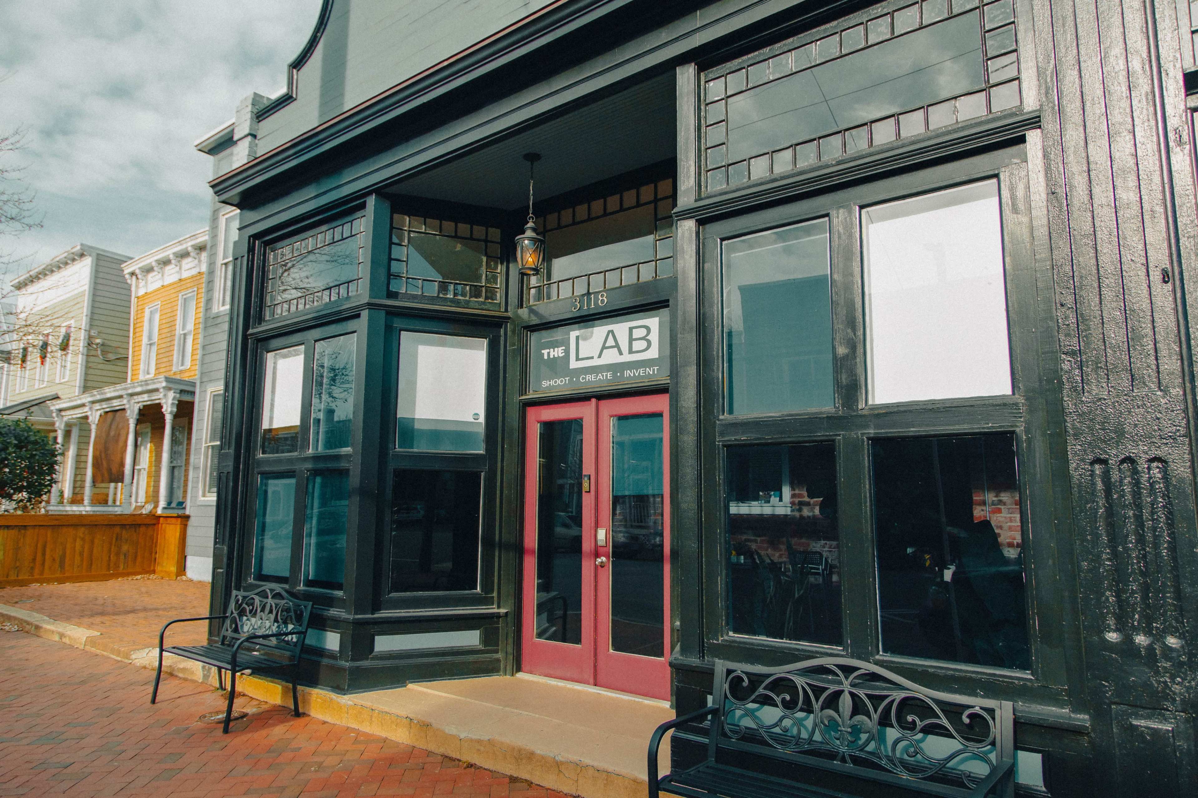 The image shows the exterior of a building with a sign that reads "The Lab," featuring large windows, a red door, and a brick sidewalk.