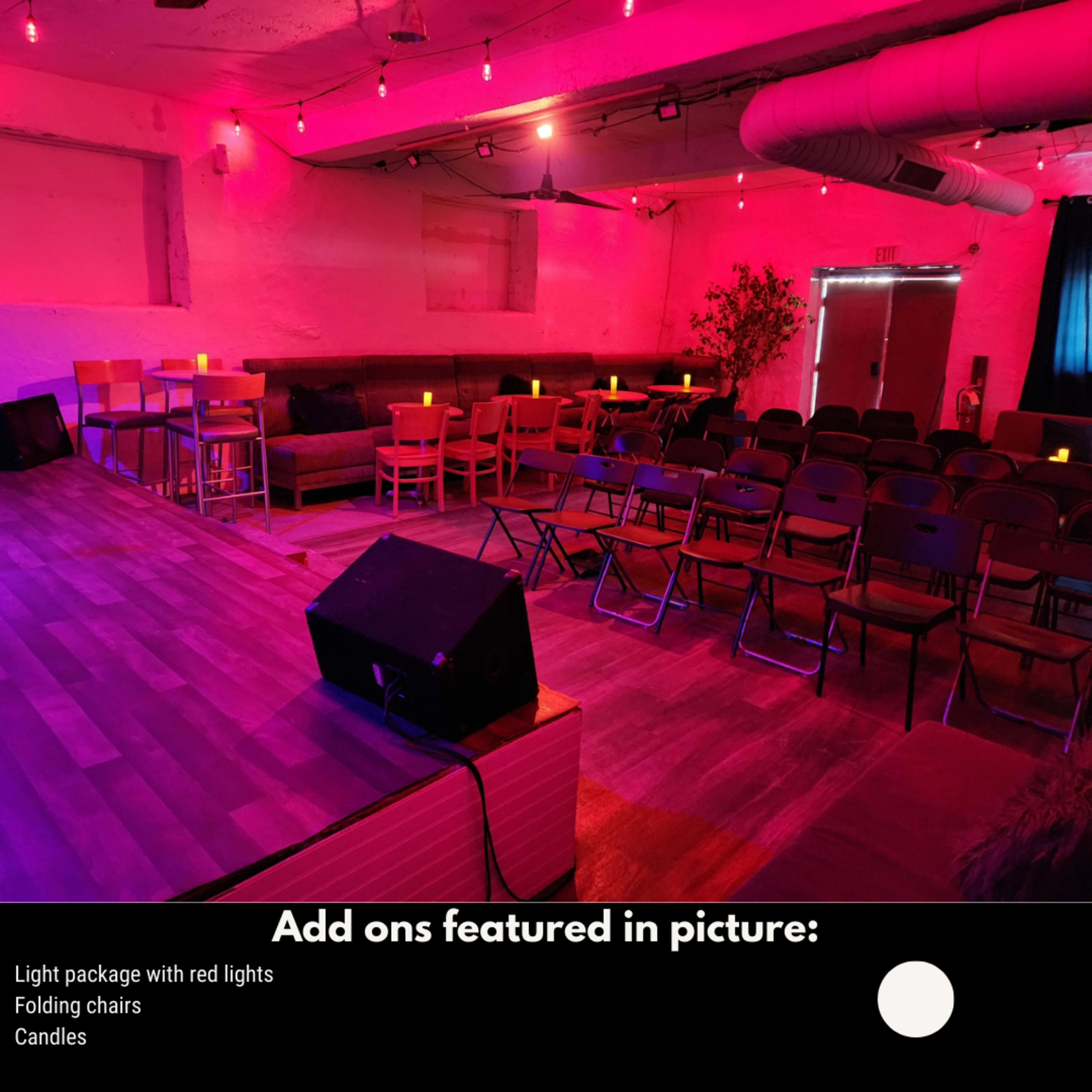 The image shows a dimly lit event space with red lights, featuring a stage area, folding chairs, rows of wooden chairs, and small candles on tables.