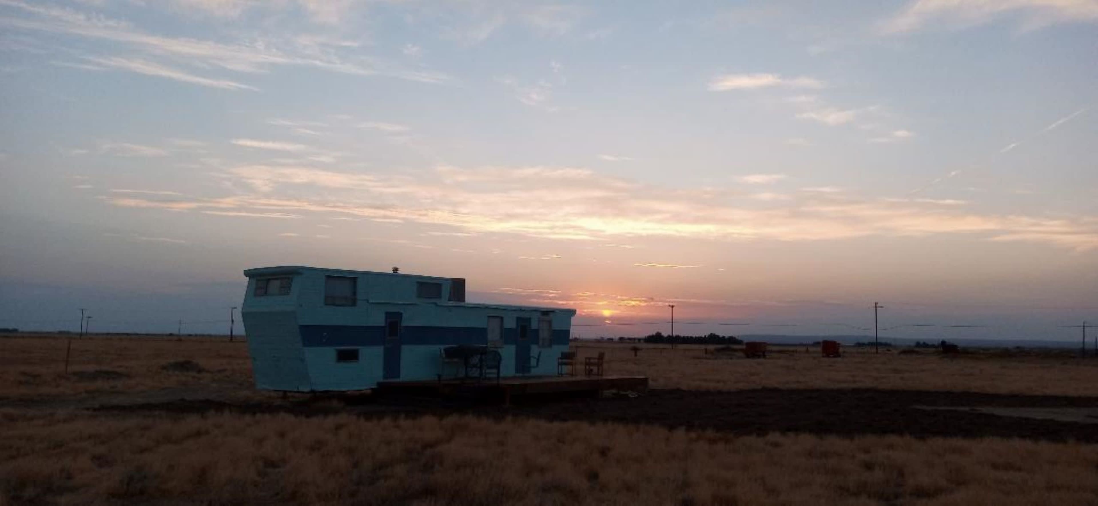 A blue mobile home stands on flat land with a sunset in the background.