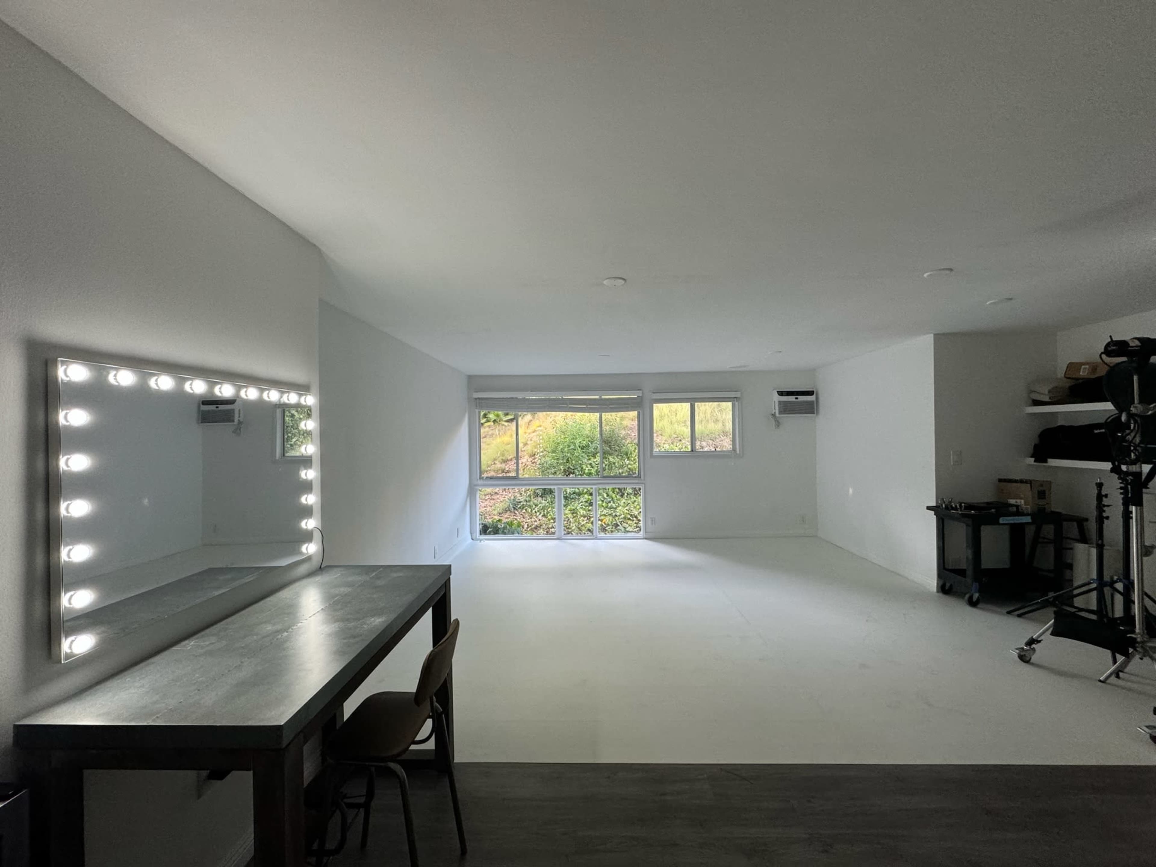 The image shows a spacious, empty room with a large mirror surrounded by lights on one wall and windows that overlook a garden area outside.