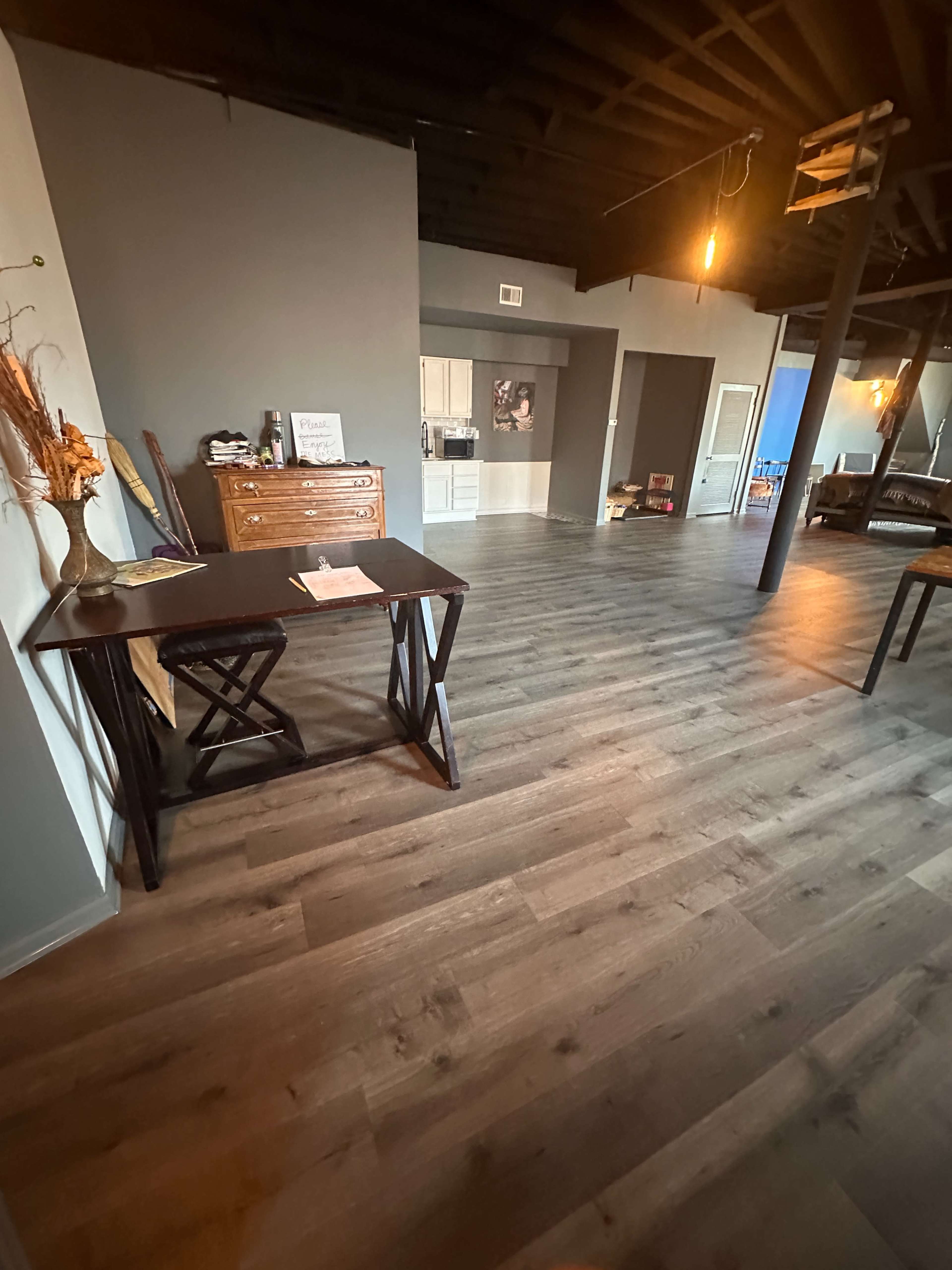 The image shows a spacious interior room with wooden flooring, a table on the left, and a kitchen area visible in the background.