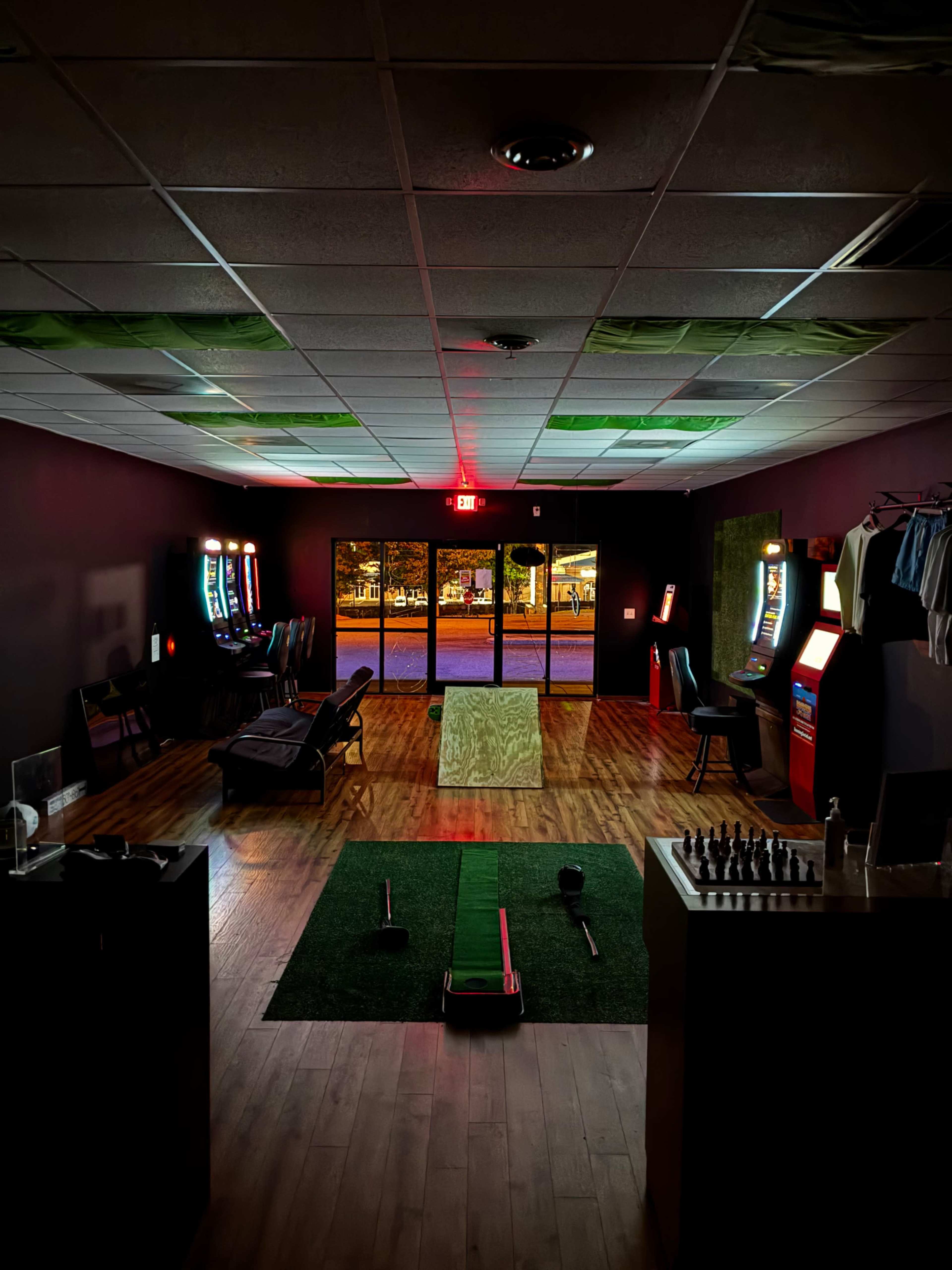 The image depicts a dimly lit entertainment space with several arcade machines, a golf putting area, and a large window showing an outdoor view at night.