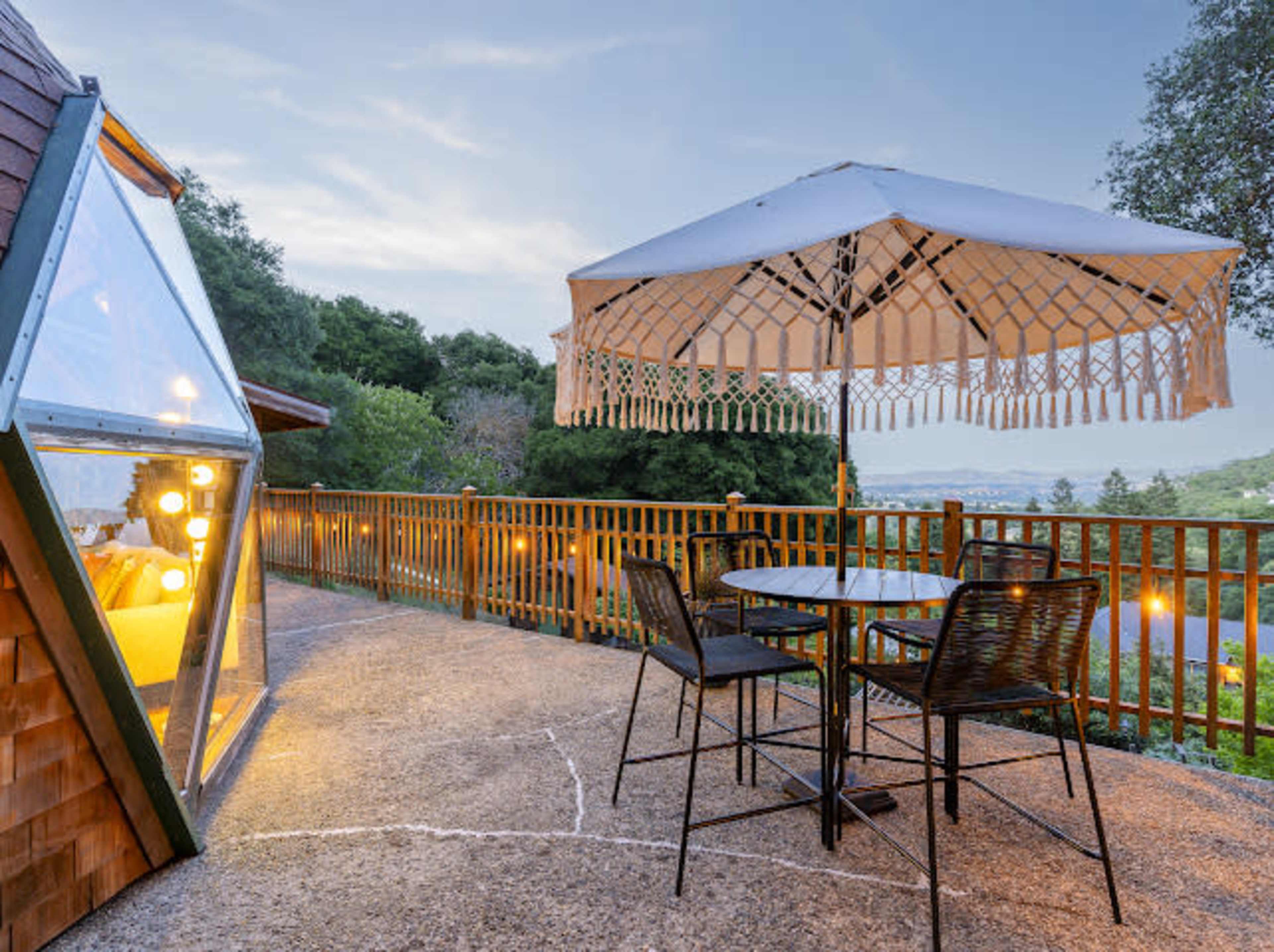 A patio area features a round table and chairs under a large umbrella, adjacent to a glass-walled structure and surrounded by lush greenery.