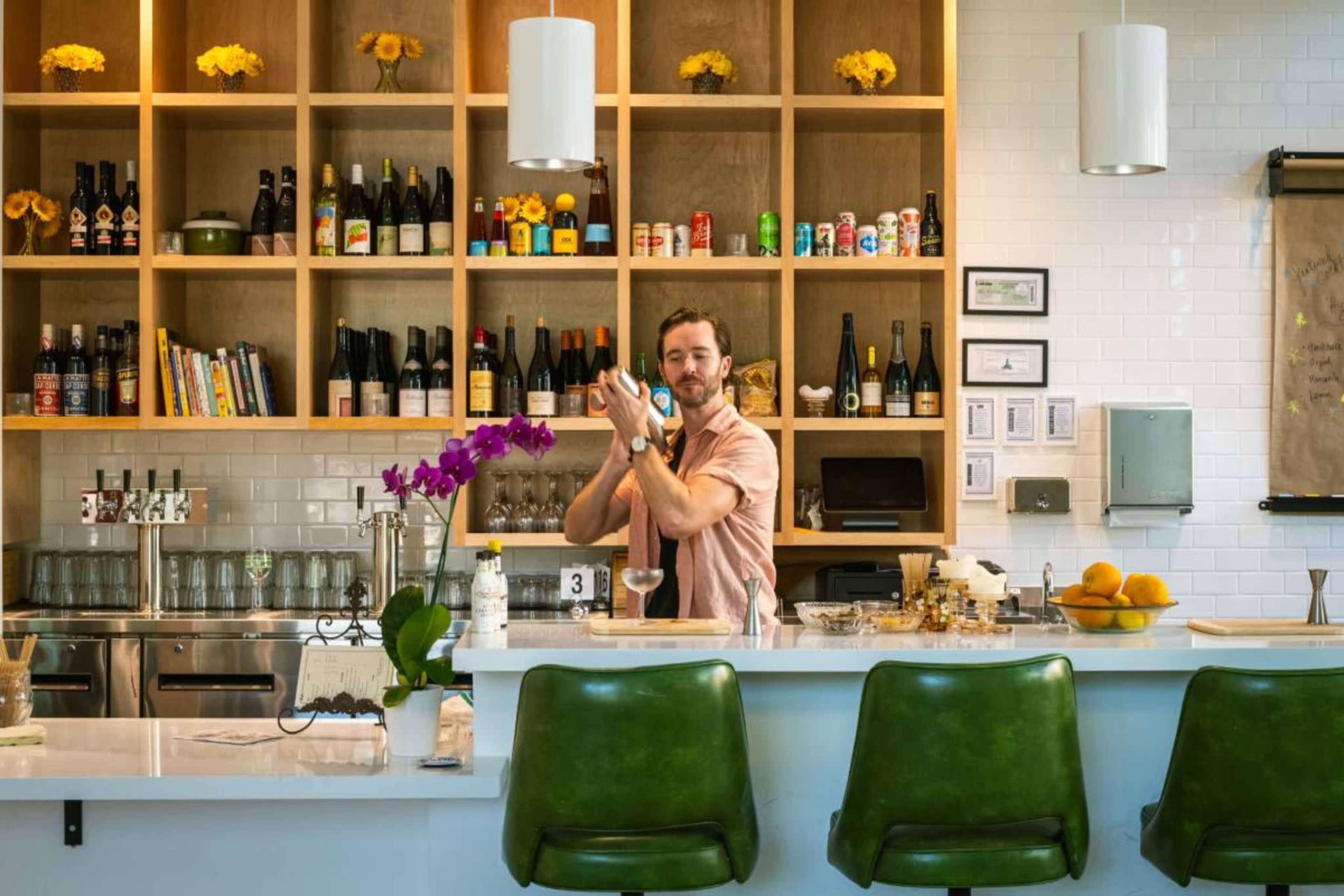 A bartender shakes a cocktail behind a well-stocked bar featuring various bottles and decorative yellow flowers.