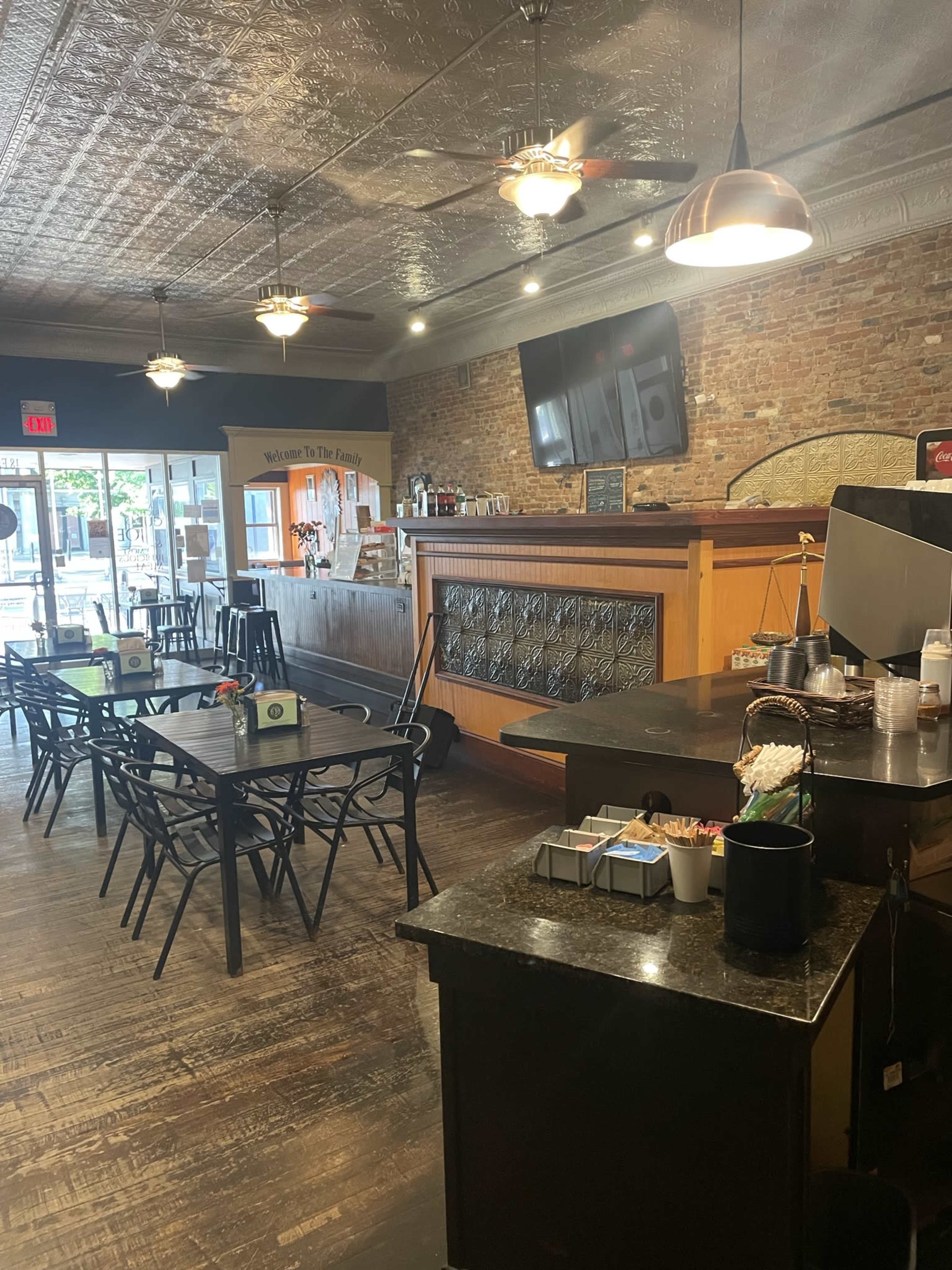 A café interior with wooden floors, tables and chairs, and a service counter featuring a large screen and various condiments.