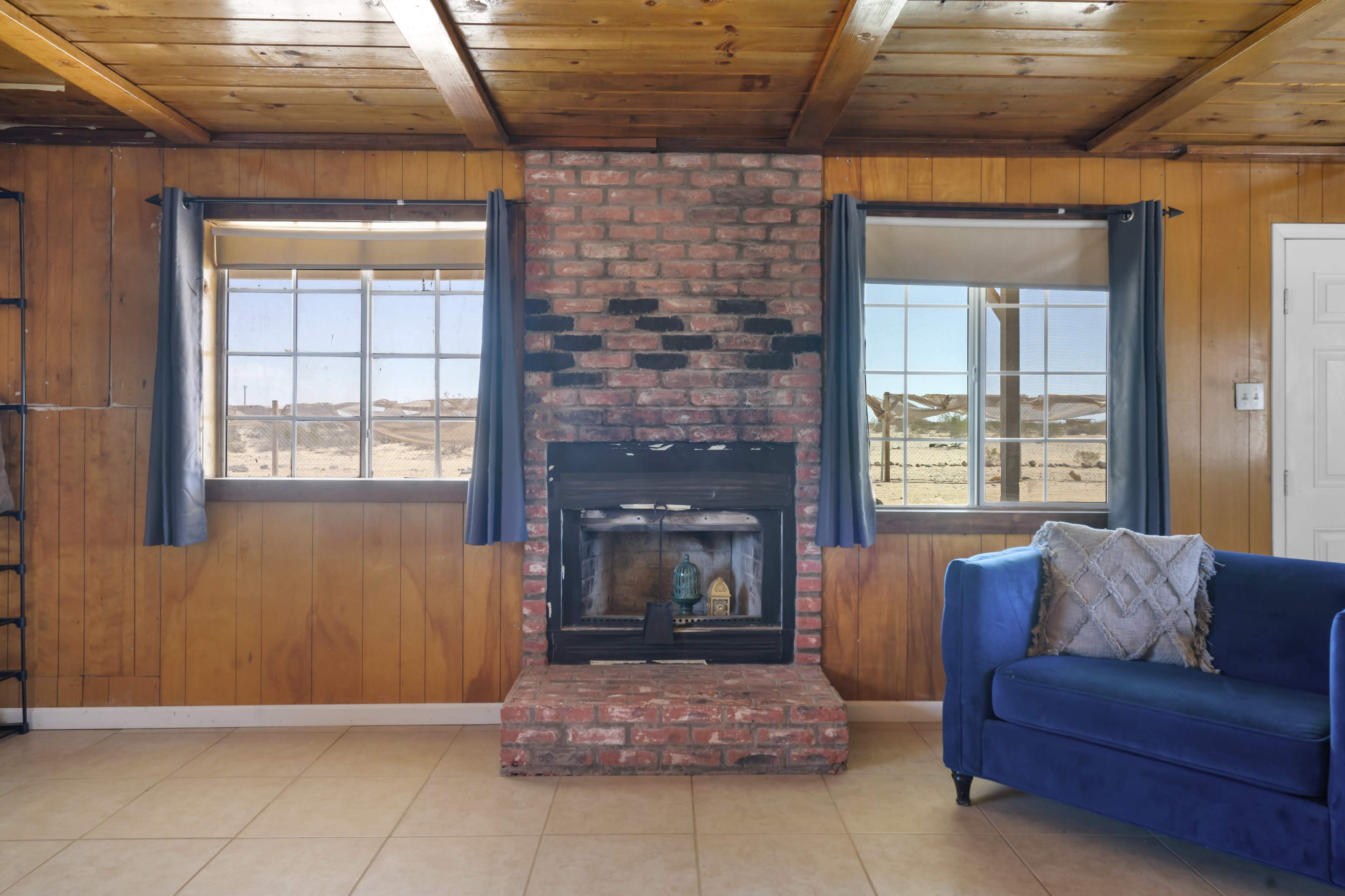A brick fireplace is centered between two windows, with a blue couch placed nearby in a room featuring wooden walls and a tiled floor.