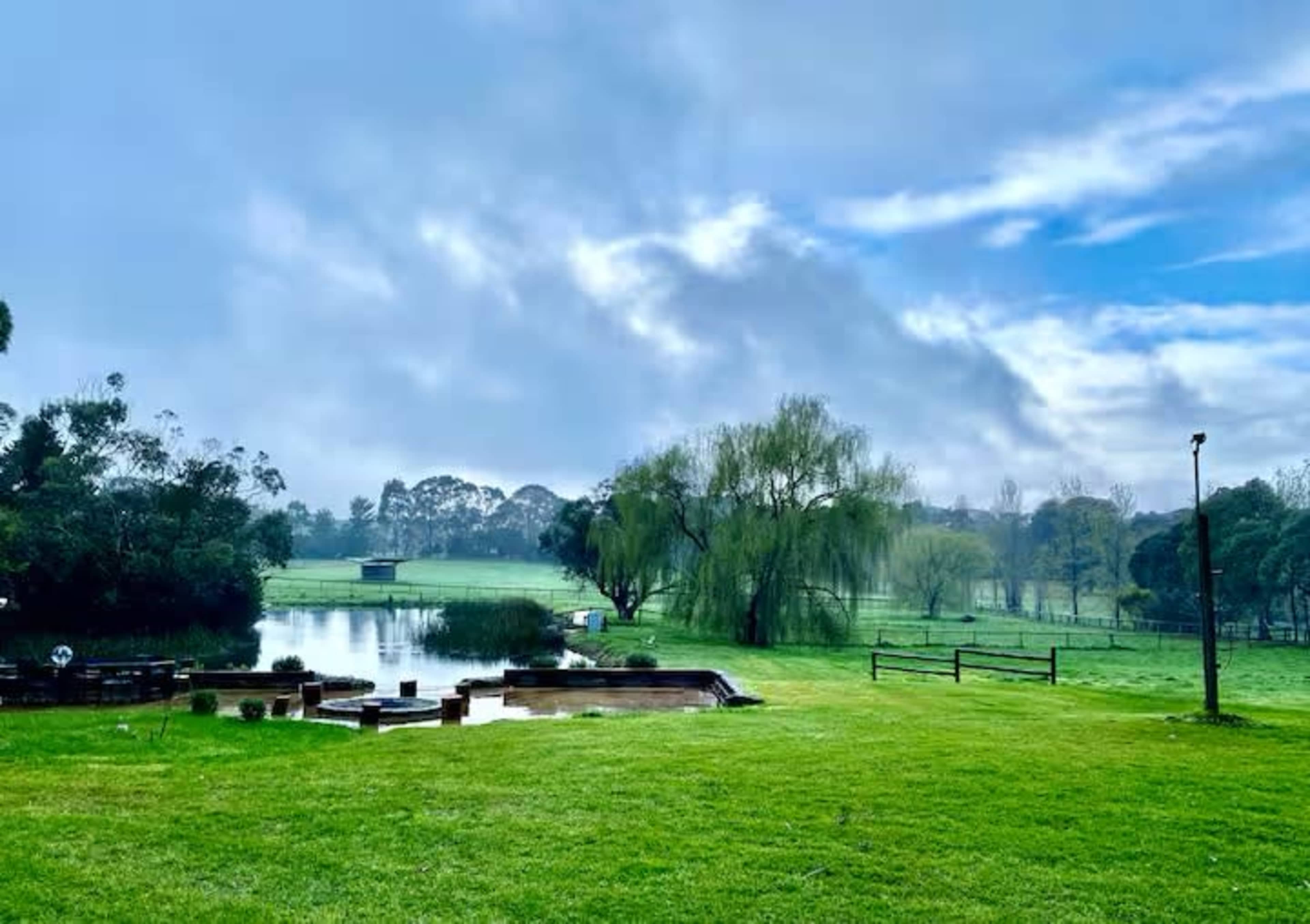 The image shows a serene landscape featuring a pond surrounded by grassy fields and trees under a cloudy sky.