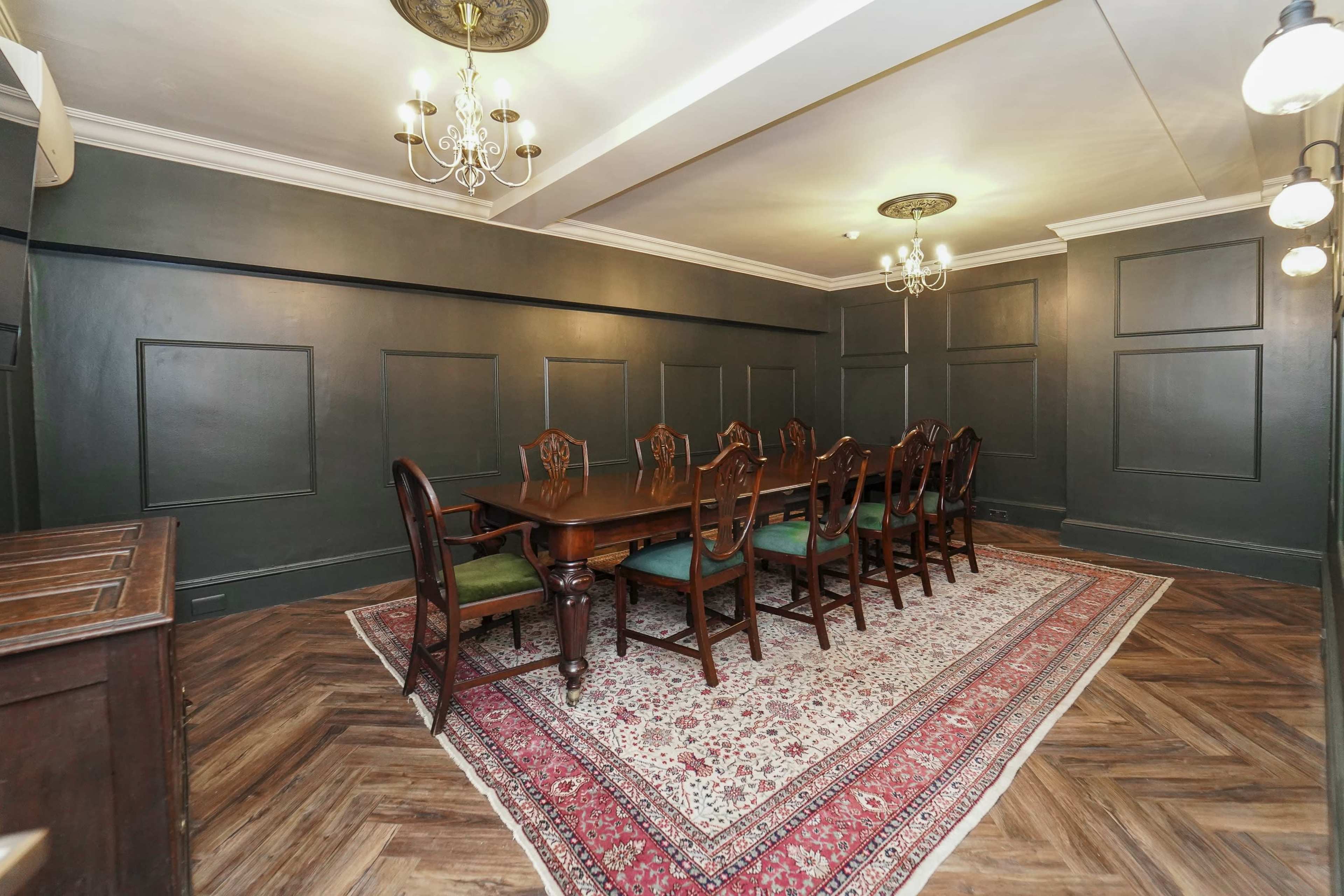 The image shows a dining room with dark green walls, a long wooden table surrounded by chairs, and a decorative rug on a herringbone-patterned floor.
