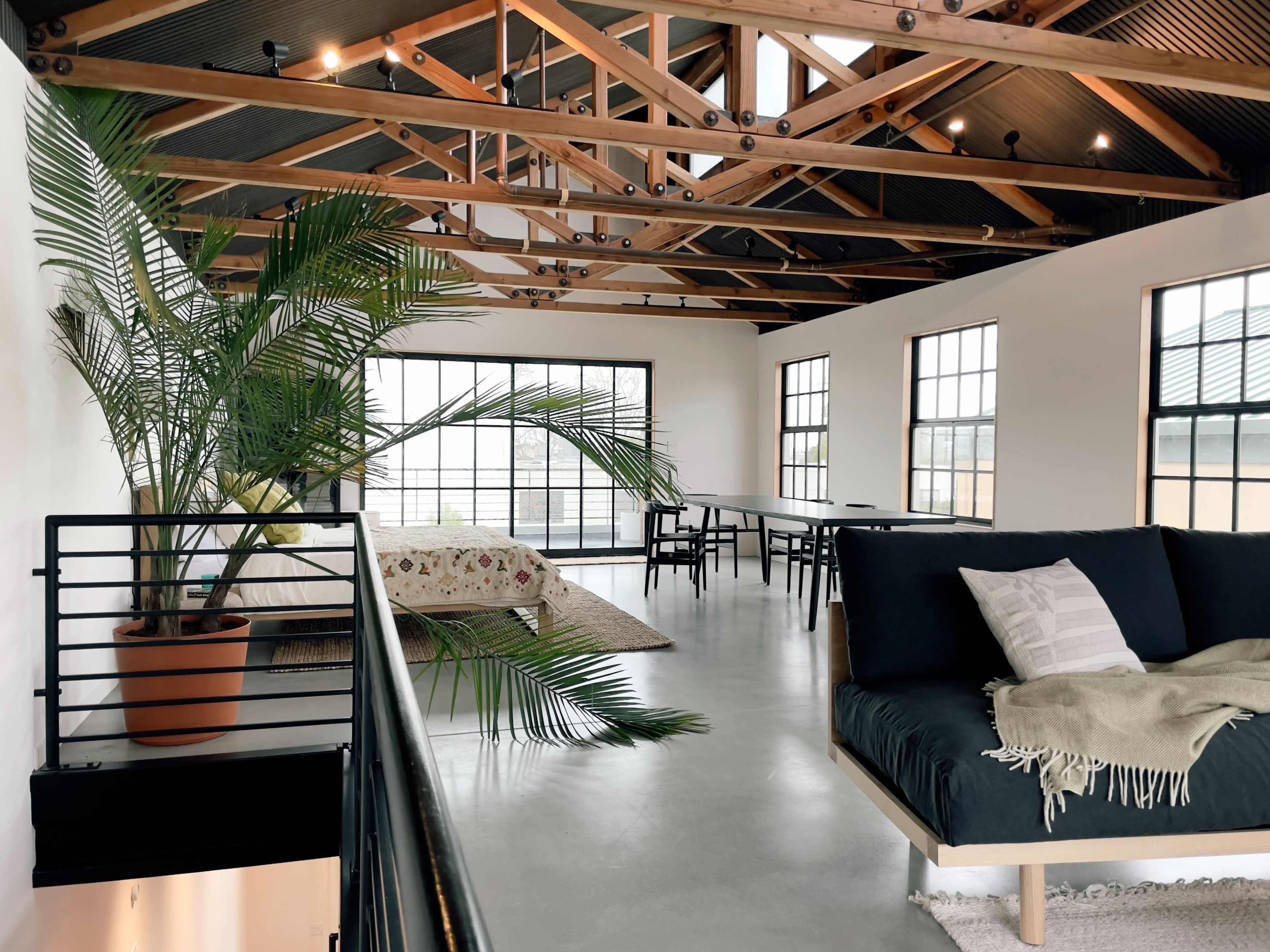A spacious, modern loft with high wooden ceilings, large windows, a sofa, a bed, and a dining area.