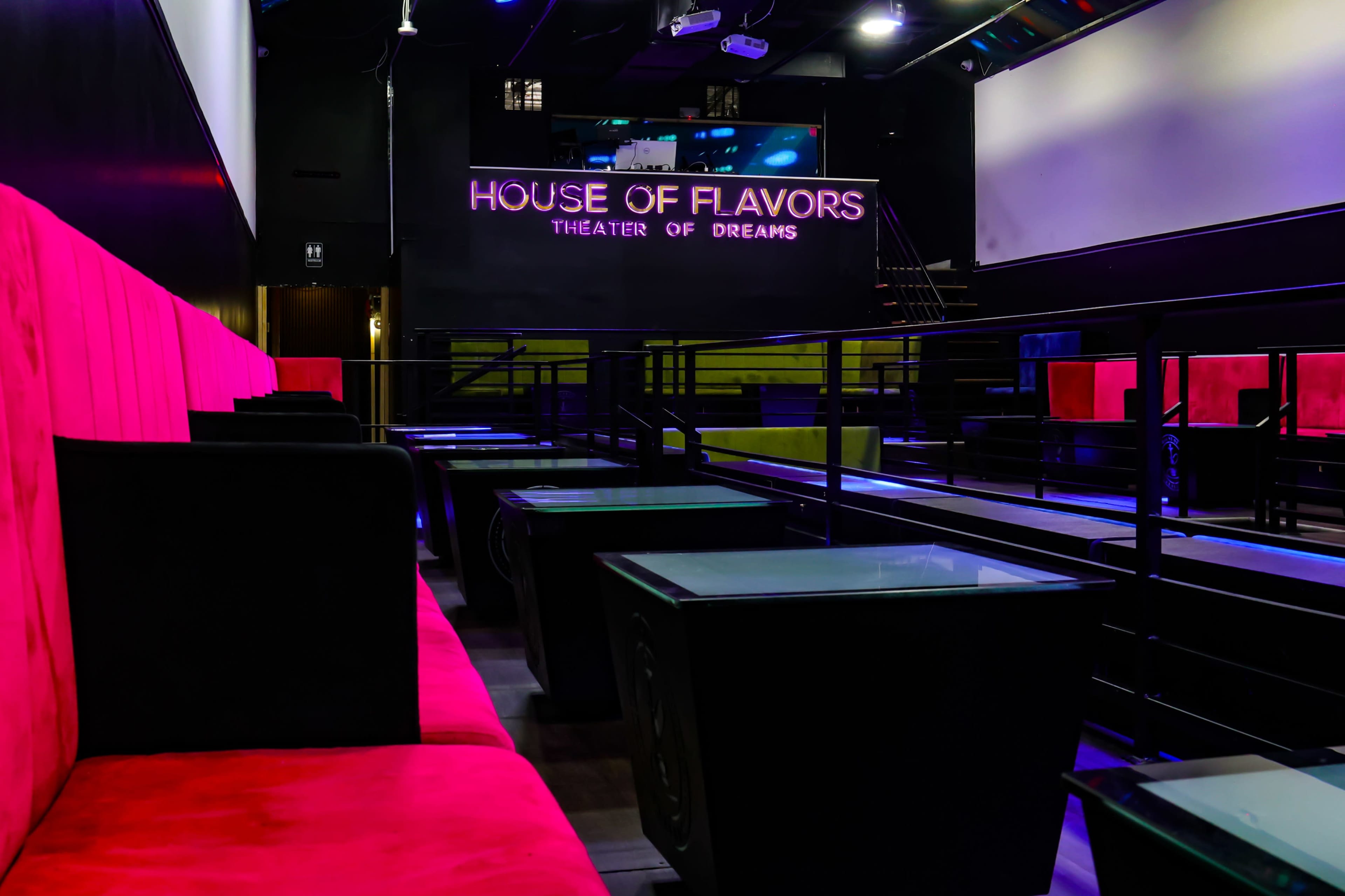 The image shows a modern lounge or theater space with pink seating and low black tables, featuring a sign that reads "HOUSE OF FLAVORS THEATER OF DREAMS" on the back wall.