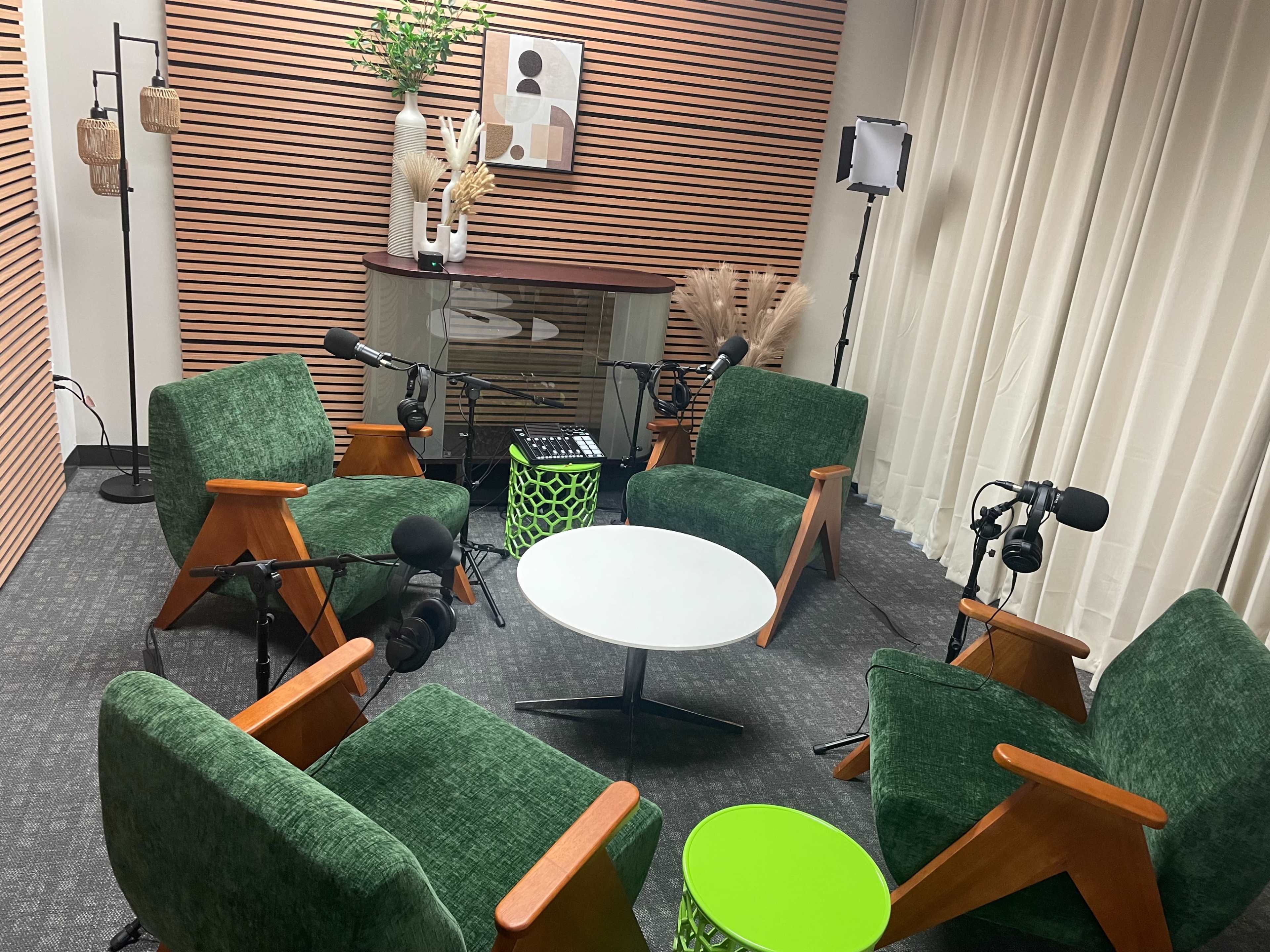 The image shows a modern recording studio with green chairs arranged around a small white table, equipped with microphones and accent decor.