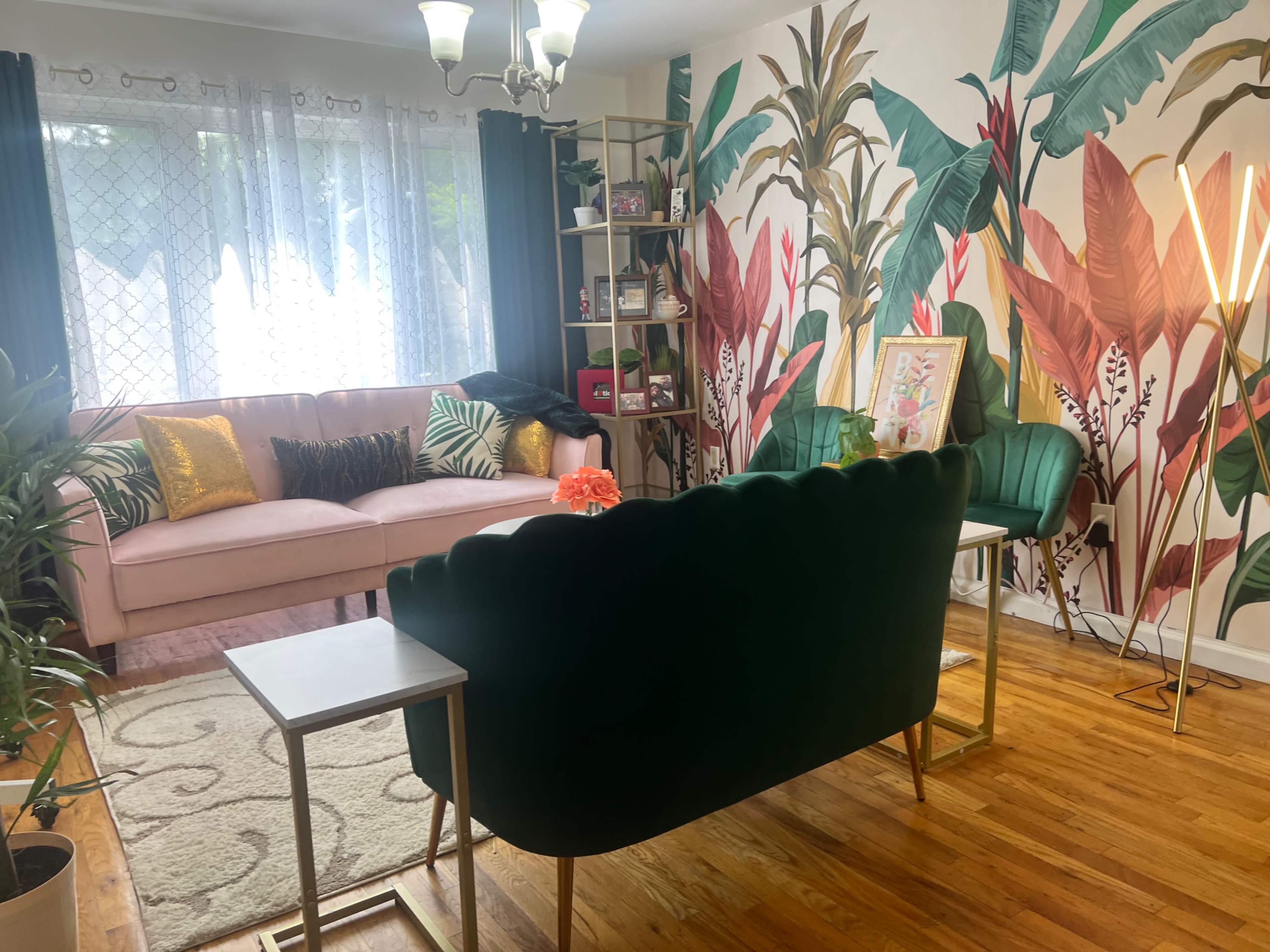 The image shows a living room featuring a pink sofa, a green velvet couch, a small white table, and a decorative wall with tropical plants wallpaper.