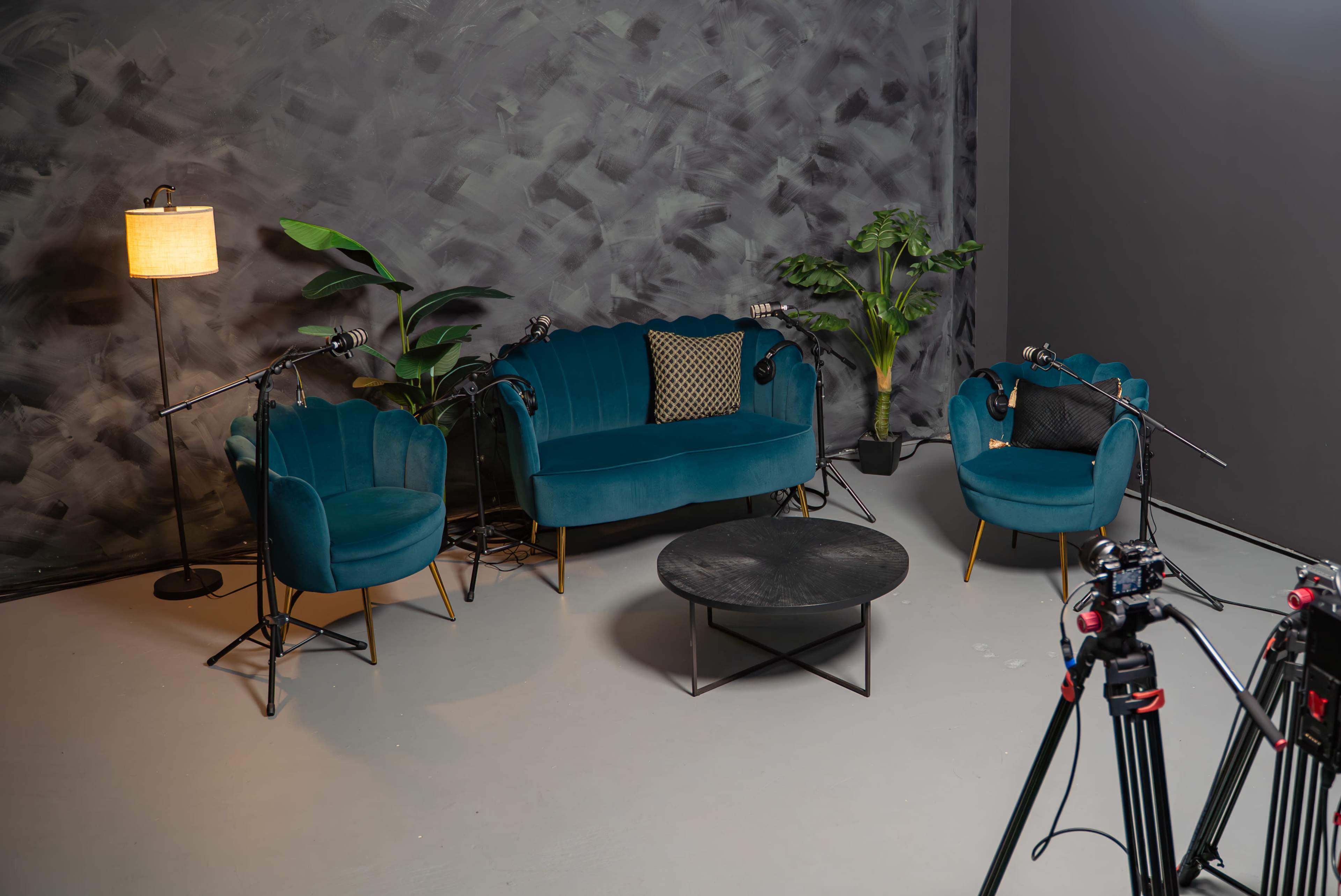 The image shows a small, modern interview set featuring two teal sofas, a round black coffee table, and a floor lamp against a textured gray wall, with several microphones positioned around the seating area.