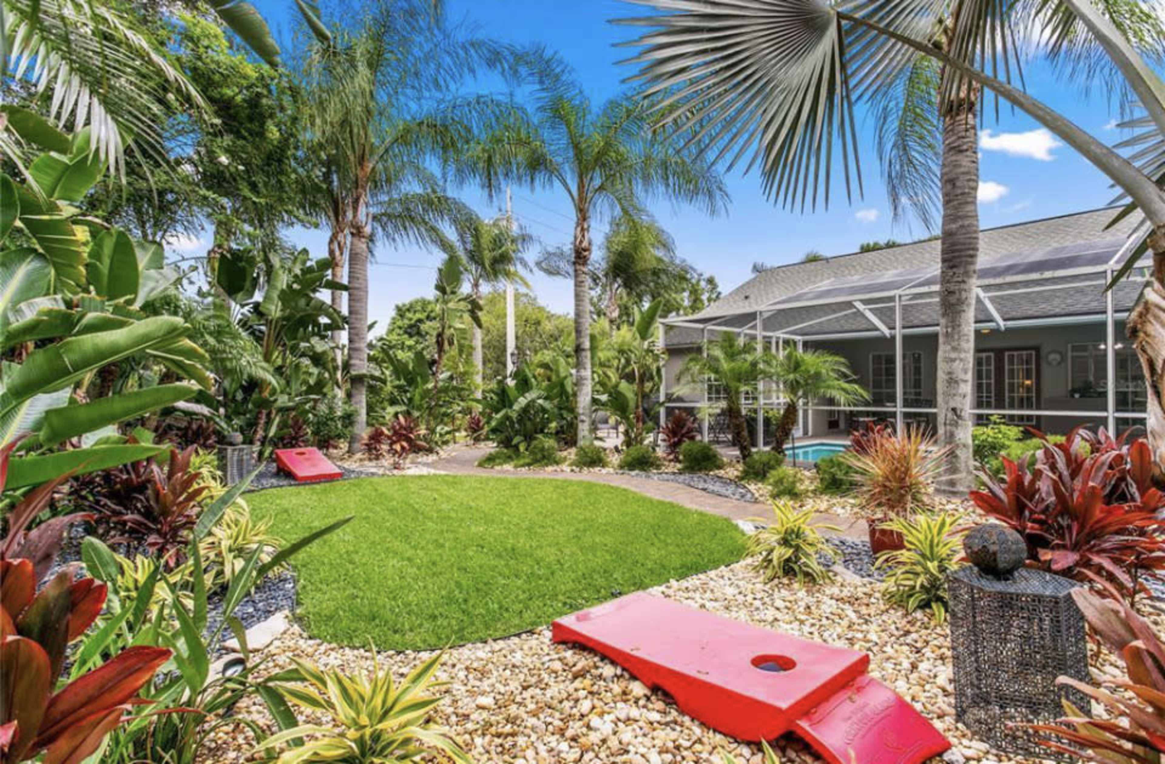 A landscaped backyard with palm trees, colorful plants, a green lawn, and cornhole boards set up on the ground.