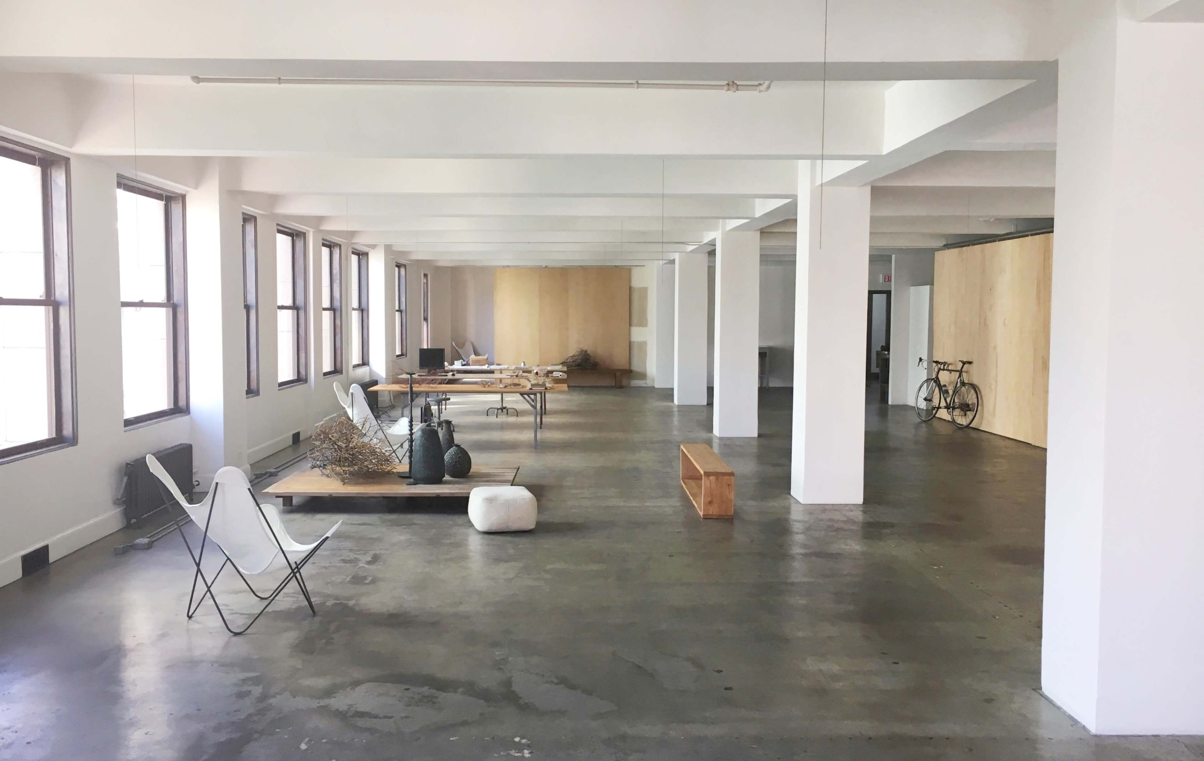 A spacious, unfinished interior with large windows, concrete floors, and scattered furniture including a table, chairs, and bicycles.