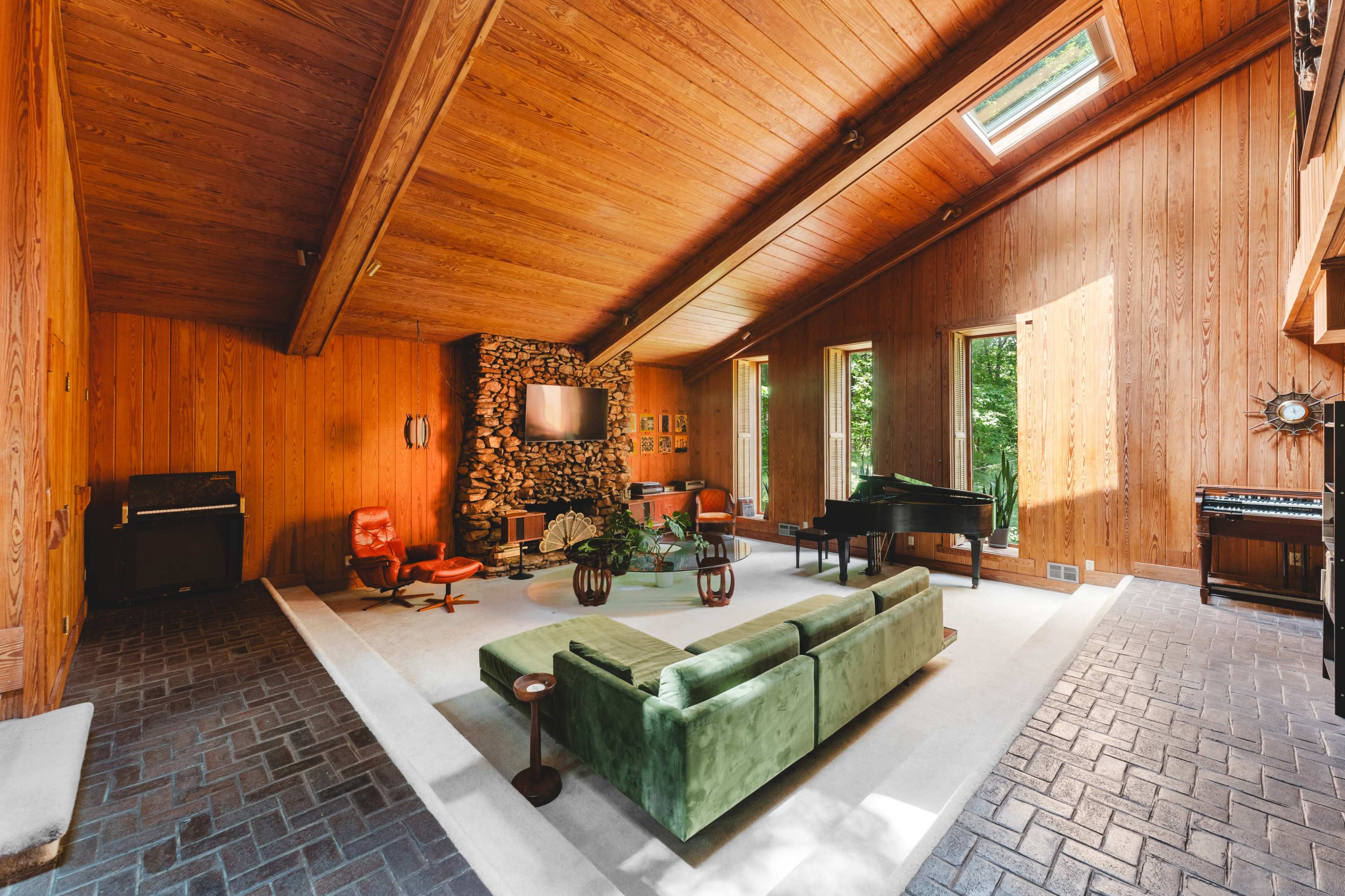 The image shows a spacious living room with wooden walls, a stone fireplace, large windows, and a mix of modern and vintage furniture, including a green sofa and a grand piano.
