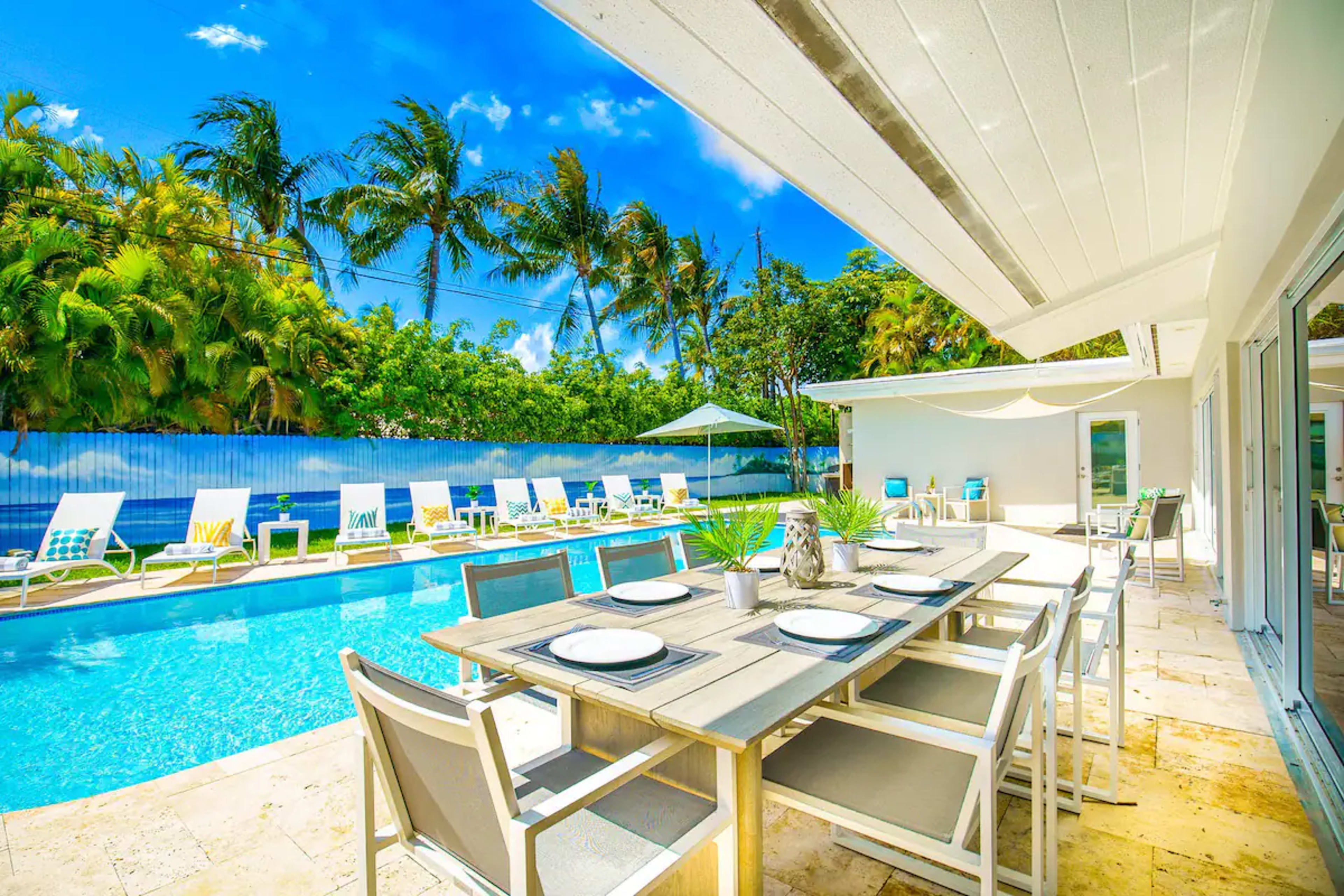 A dining table is set near a swimming pool surrounded by palm trees and lounge chairs.