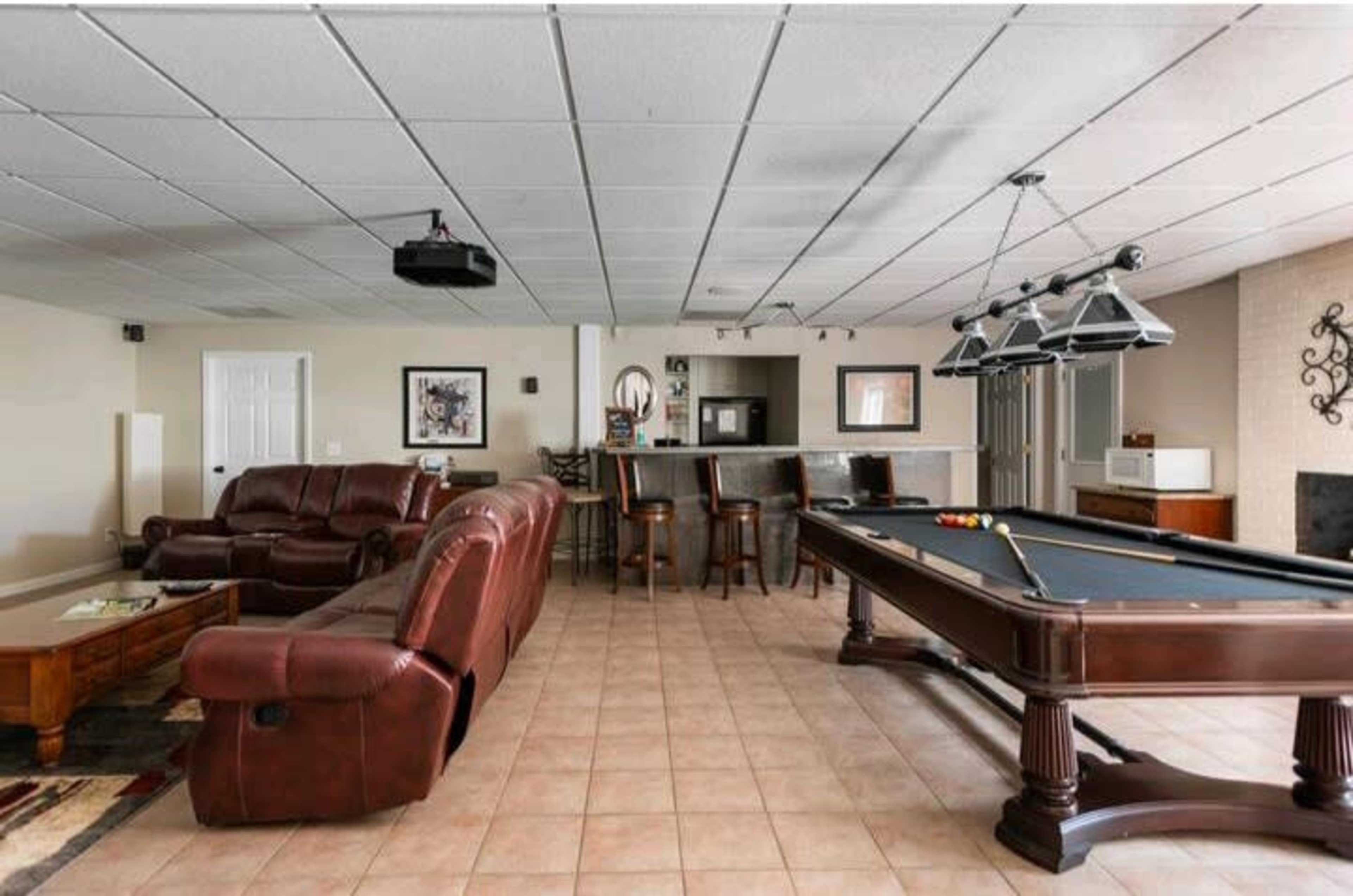 A spacious recreational room featuring a pool table, a seating area with leather couches, and a bar counter with high stools.