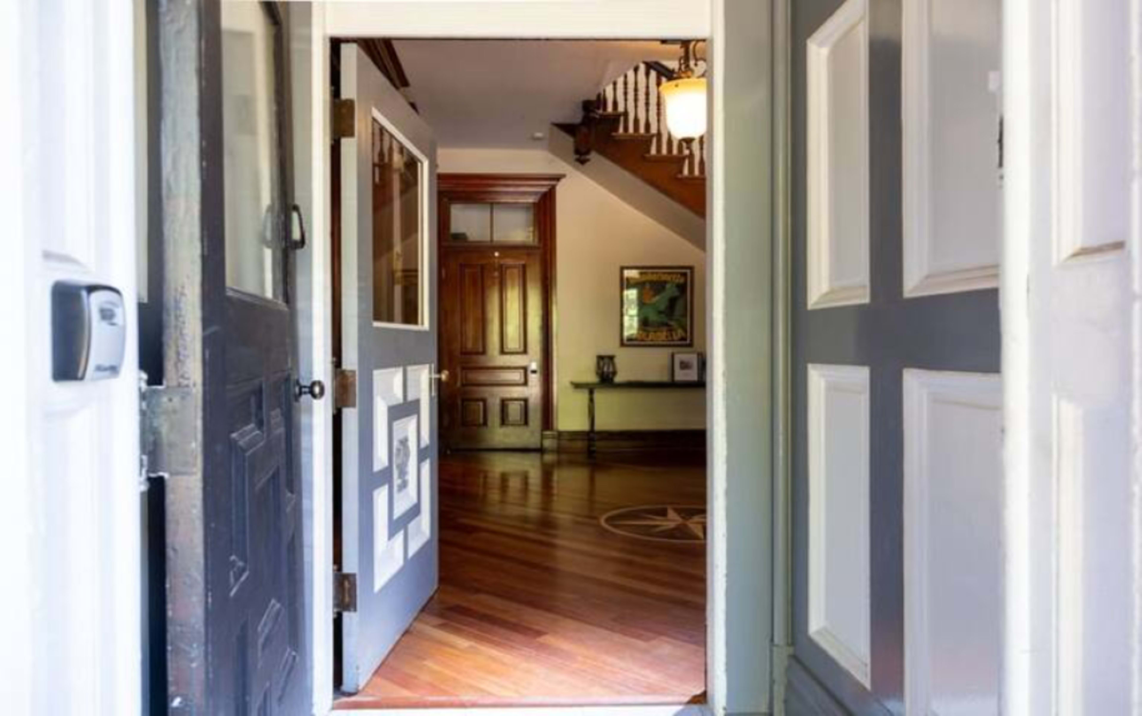The image shows an entrance hallway with an open double door leading into a wooden-floored interior space featuring a staircase and artwork on the walls.