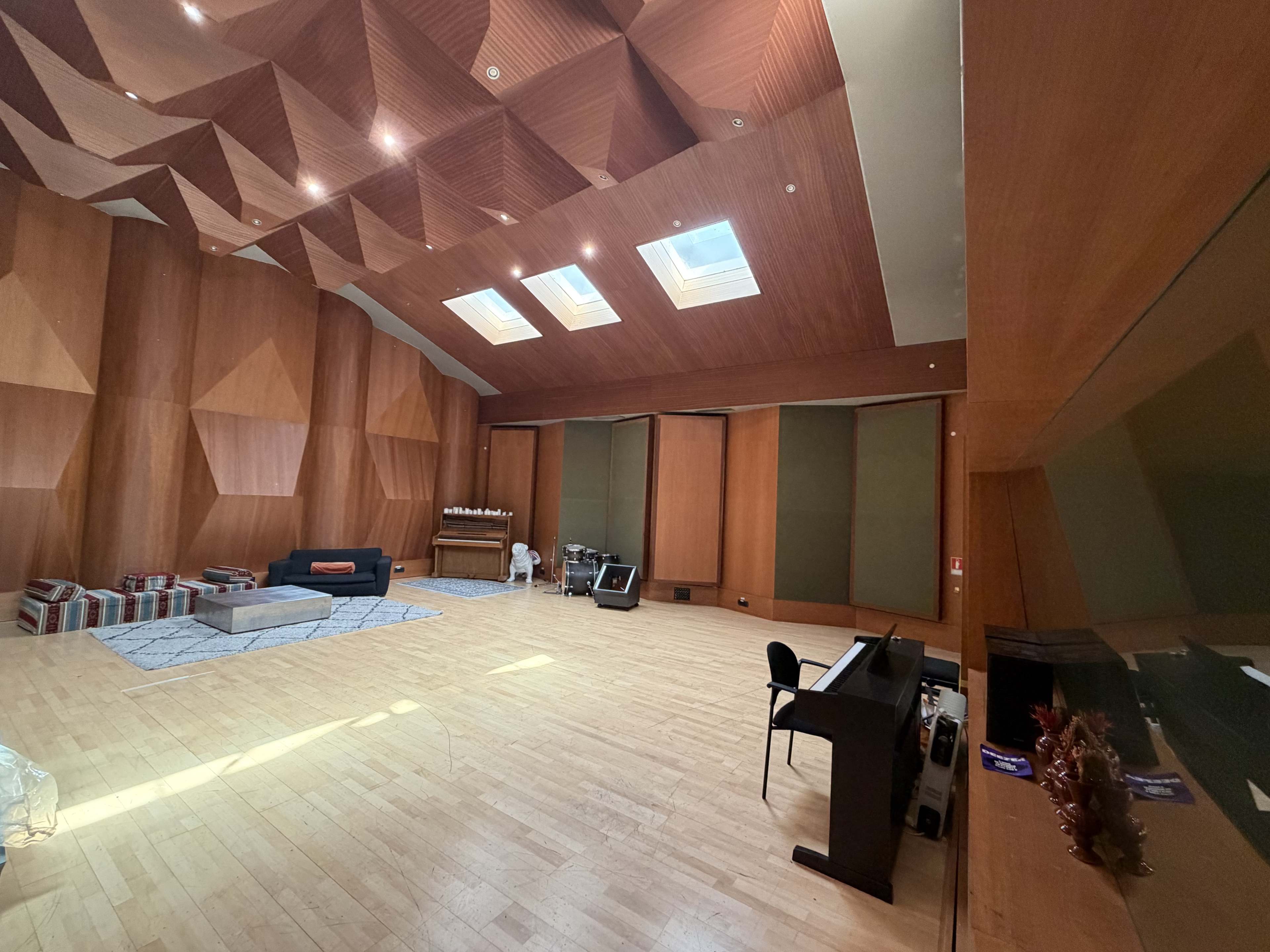 The image shows a spacious room with wooden walls, a patterned ceiling, a piano, and minimal furnishings, including a couch and a rug on the wooden floor.