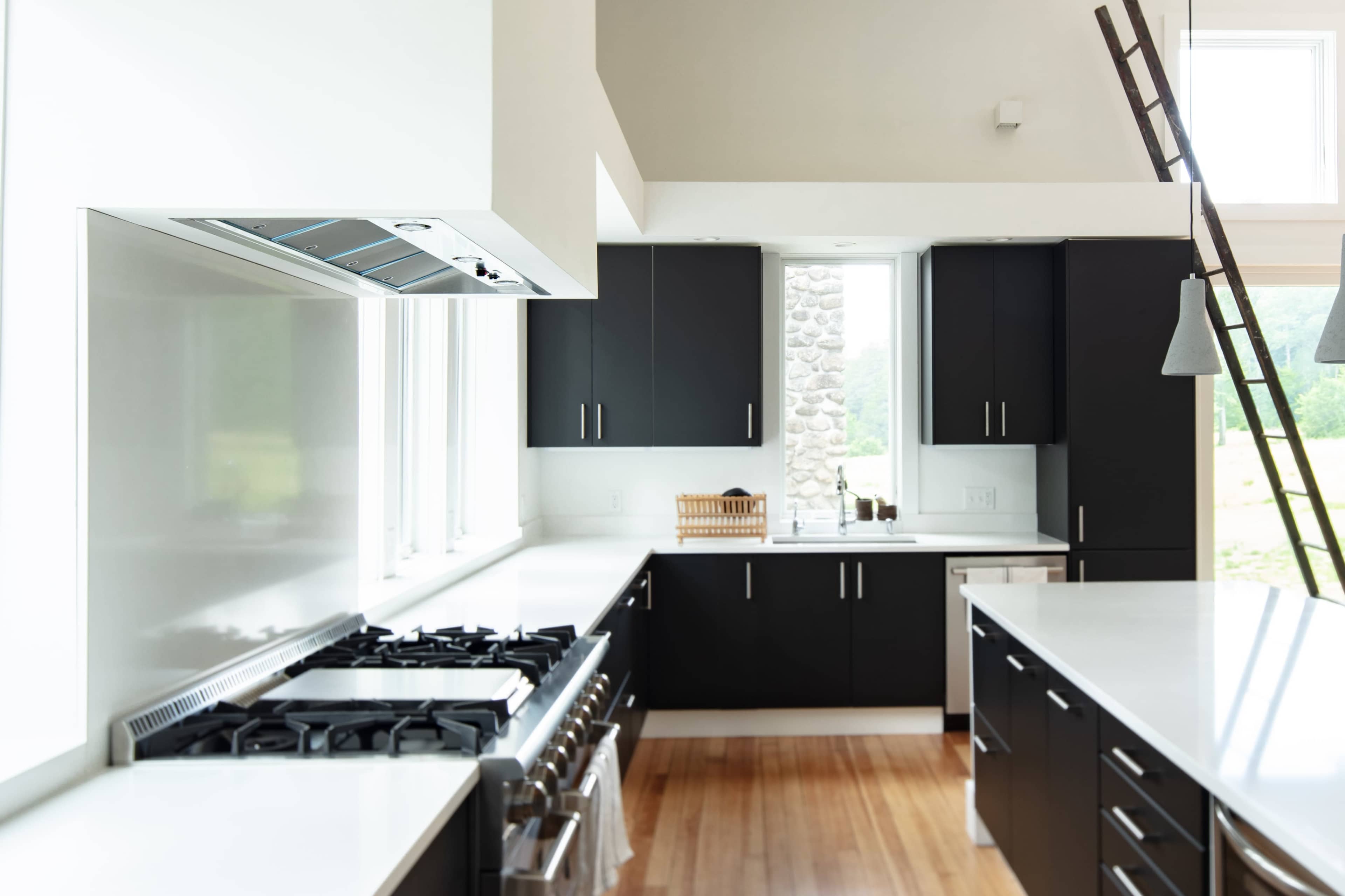 A modern kitchen features black cabinets, a stainless steel range, and large windows overlooking a natural landscape.
