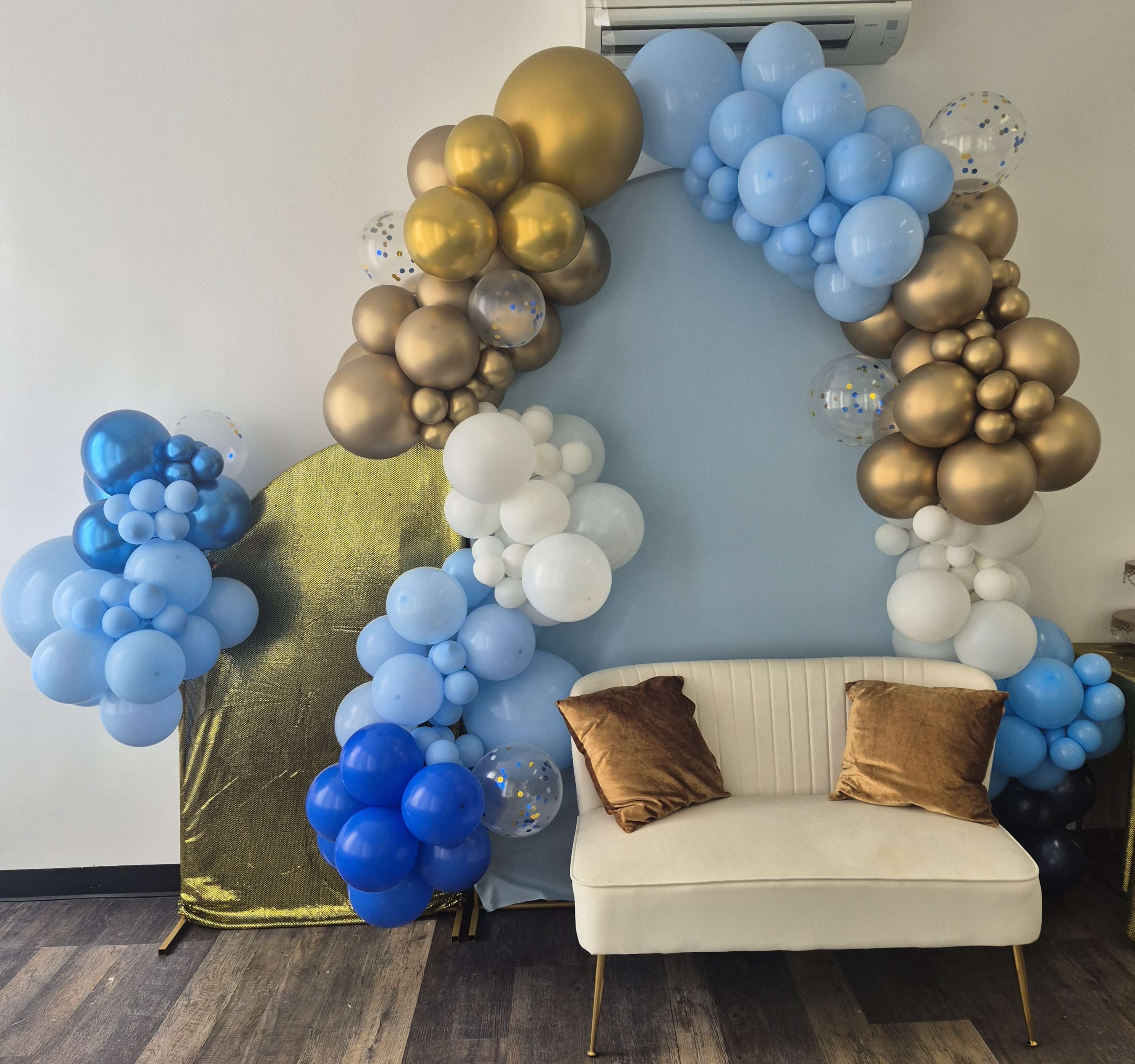 The image features a balloon arch decorated with blue, white, and gold balloons, adjacent to a gold backdrop and a white couch with two brown cushions.