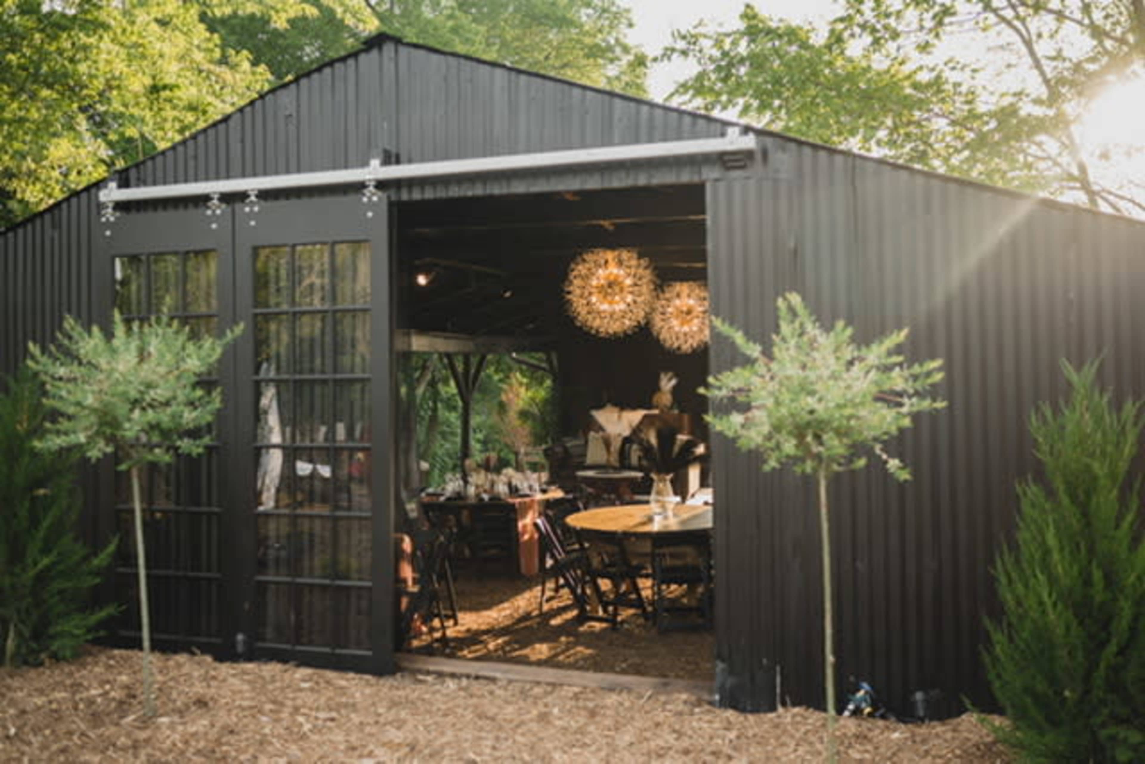 A black corrugated metal building with large glass doors opens to reveal a dining area adorned with round chandeliers and wooden tables, surrounded by greenery.