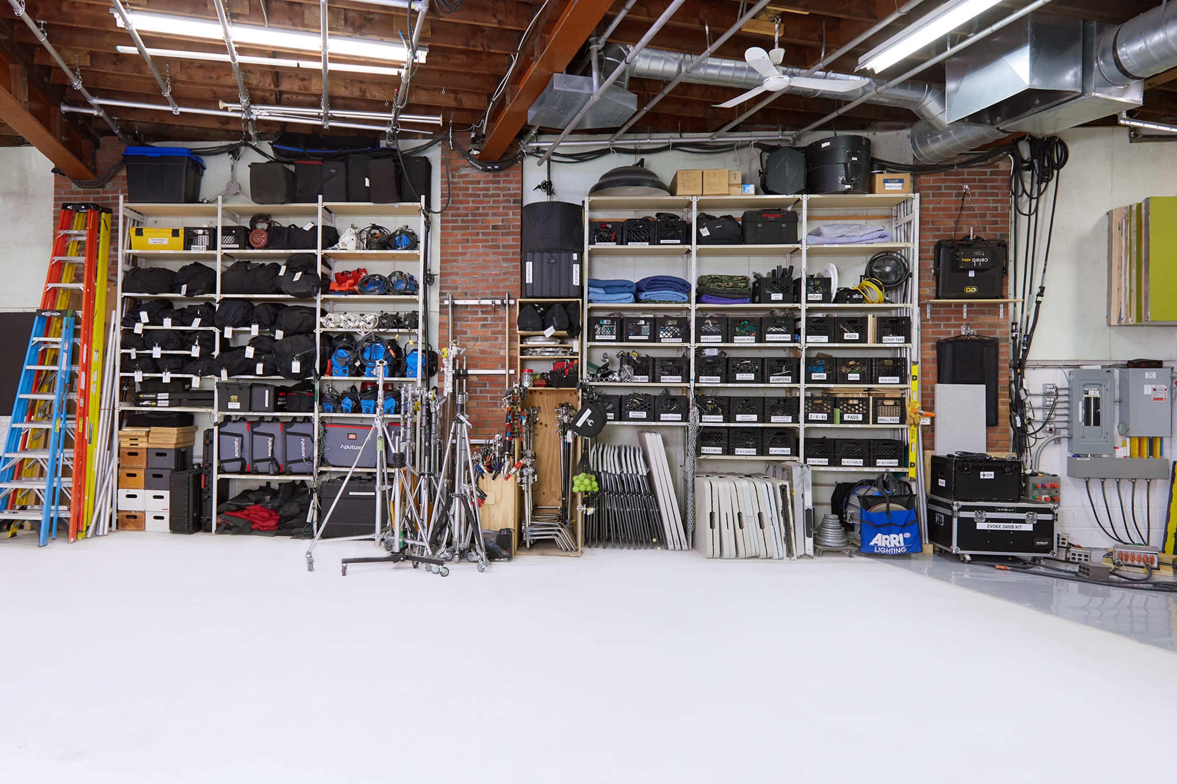 The image depicts a neatly organized storage area filled with various equipment and supplies, including shelving units, containers, and ladders.