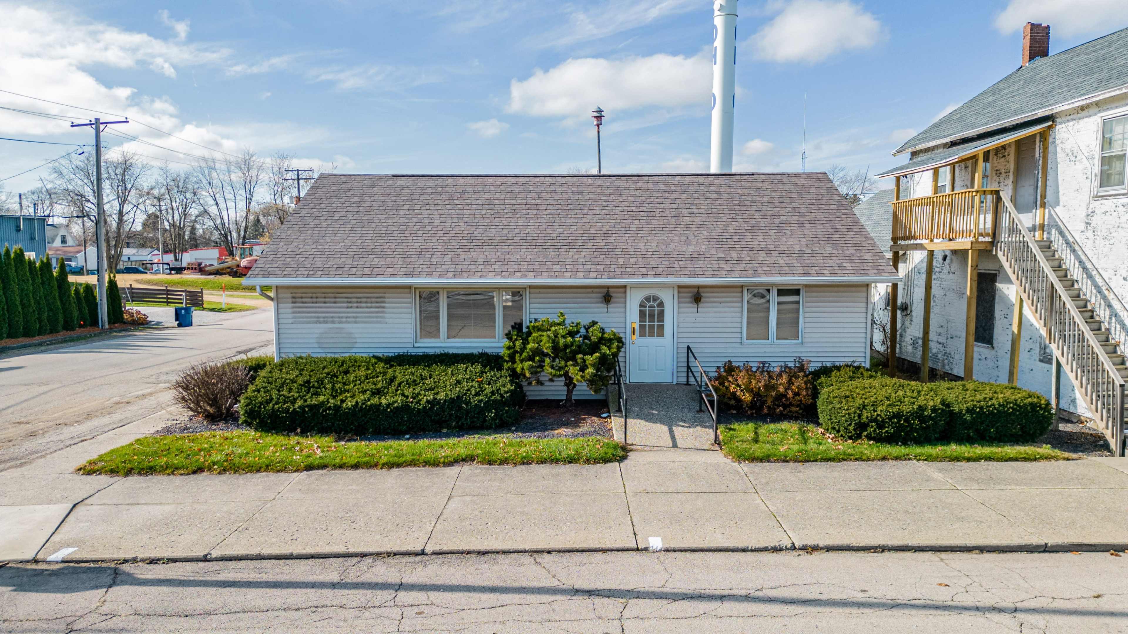 A single-story white house with a sloped roof and a front walkway is situated on a street lined with hedges and nearby buildings.