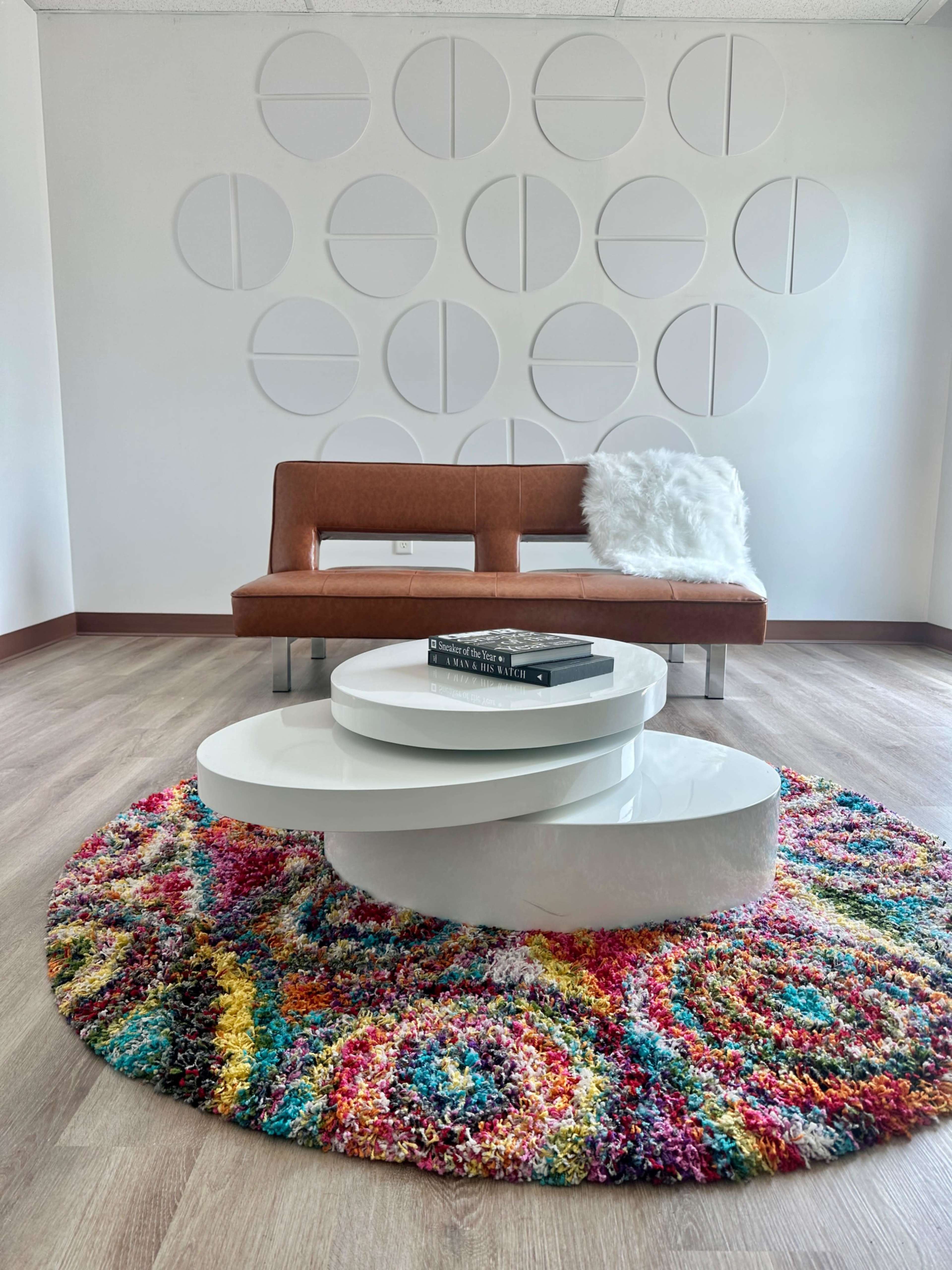 A modern seating area features a brown sofa, a round colorful rug, and a white coffee table, with a textured wall in the background.