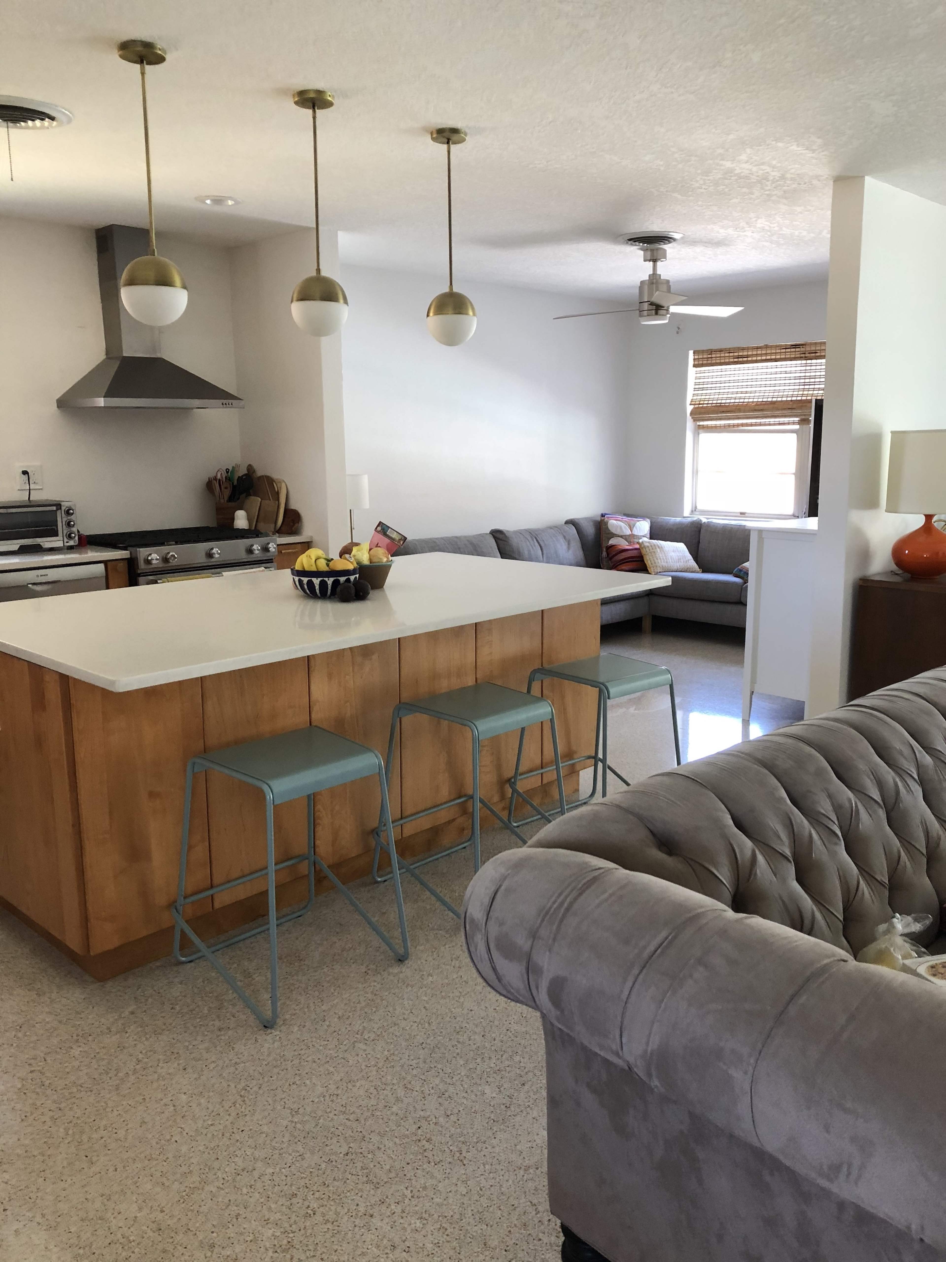 The image shows a modern kitchen with a central island and bar stools, connected to a living area furnished with a gray sofa.