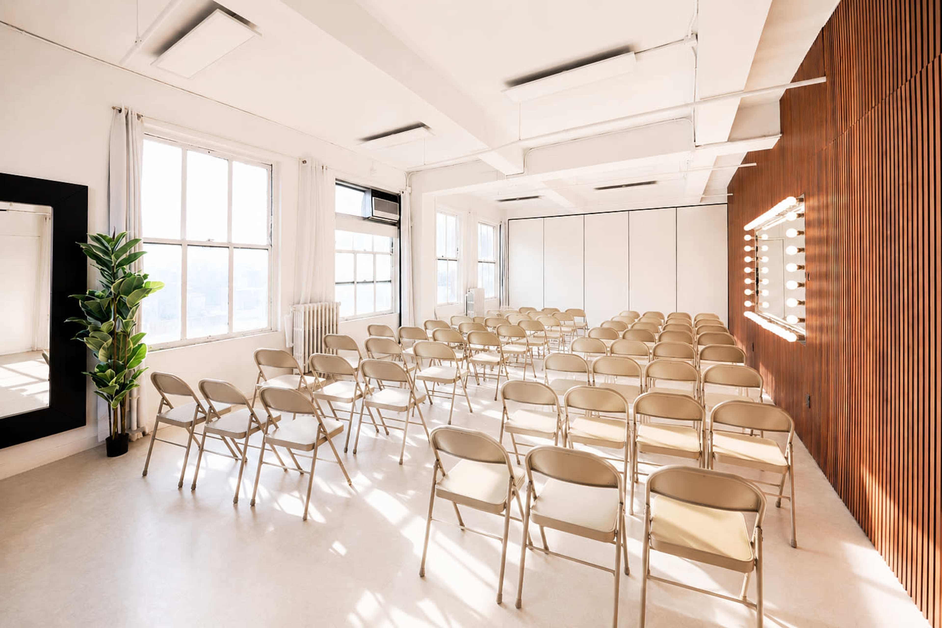 The image shows a bright, empty room set up with rows of beige folding chairs facing a mirrored wall with lights.