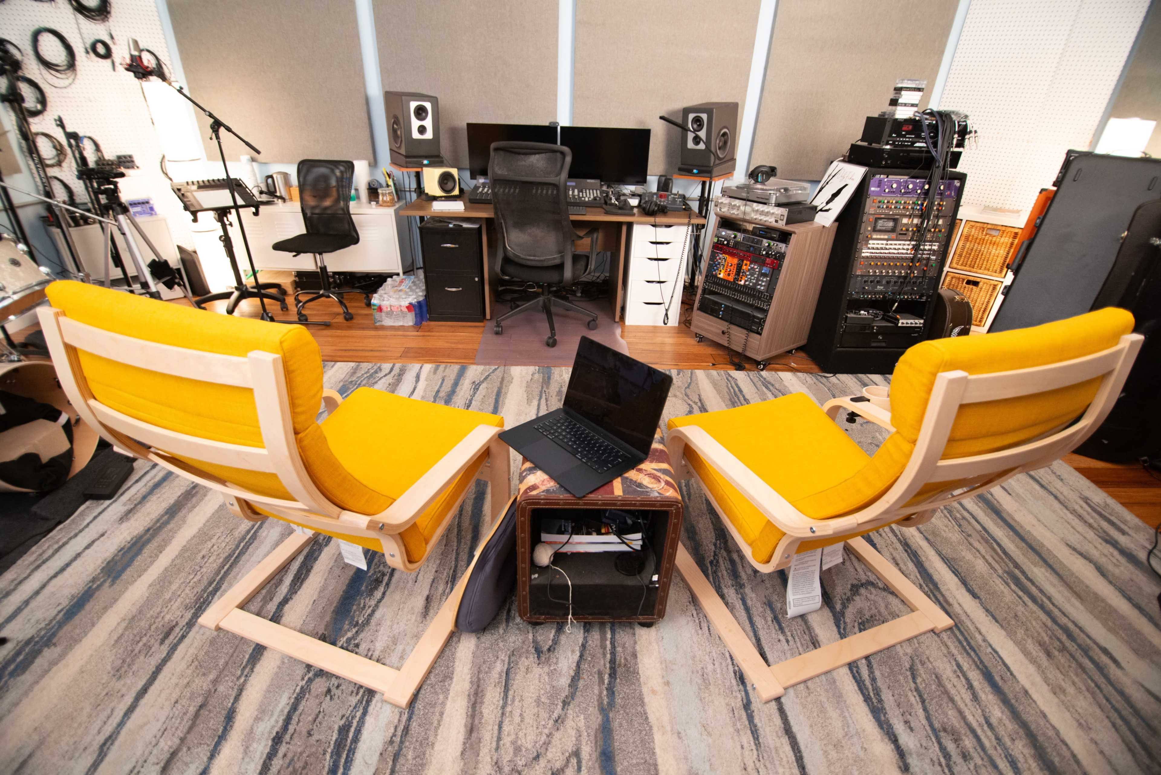 The image shows a modern music studio with two yellow lounge chairs facing a small table, and various sound equipment and instruments in the background.