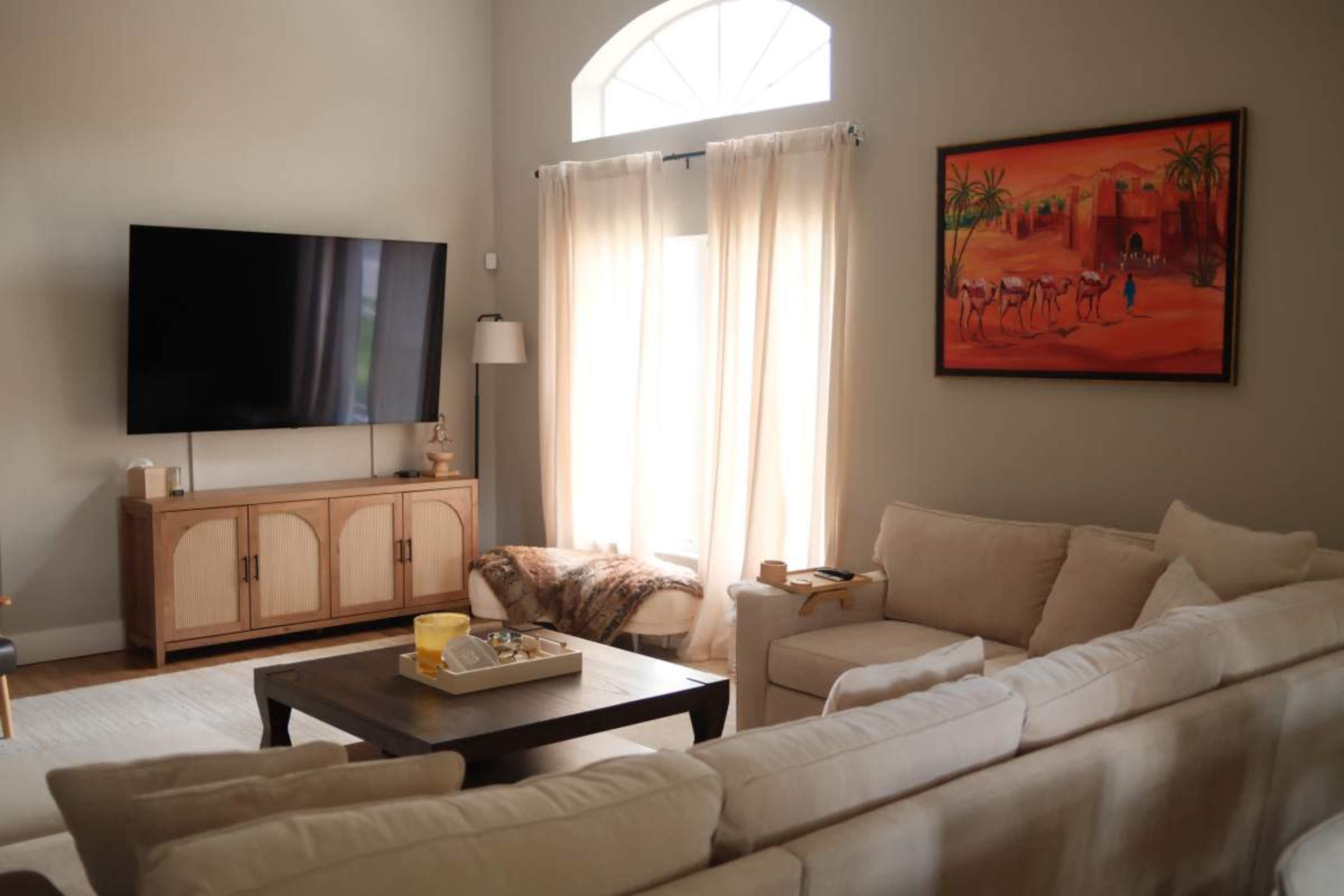 A bright living room features a large TV on a wooden cabinet, a beige sectional sofa, a coffee table, and a colorful painting on the wall.