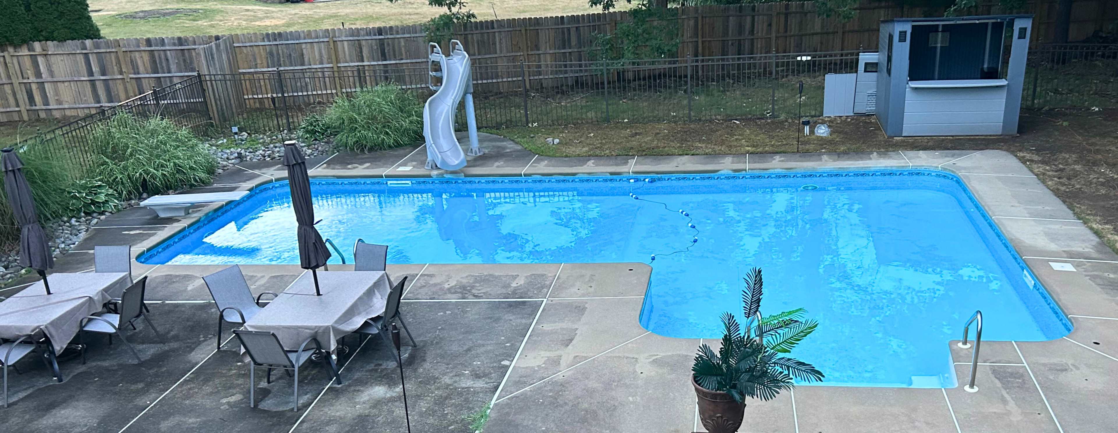 The image shows a backyard swimming pool with a slide, surrounded by patio furniture and a small outdoor kitchen area.